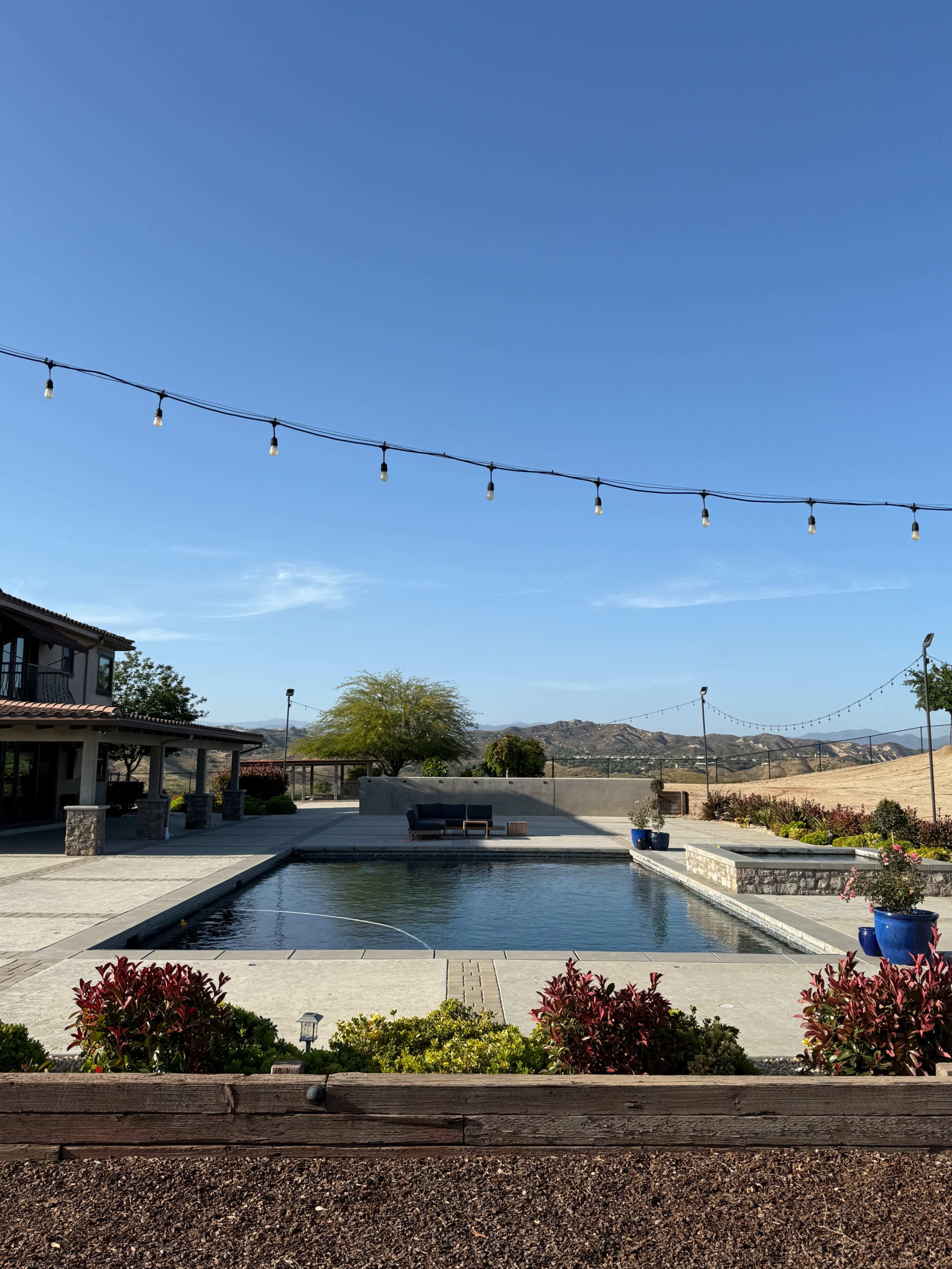 A swimming pool surrounded by landscaped plants and distant hills under a clear blue sky.