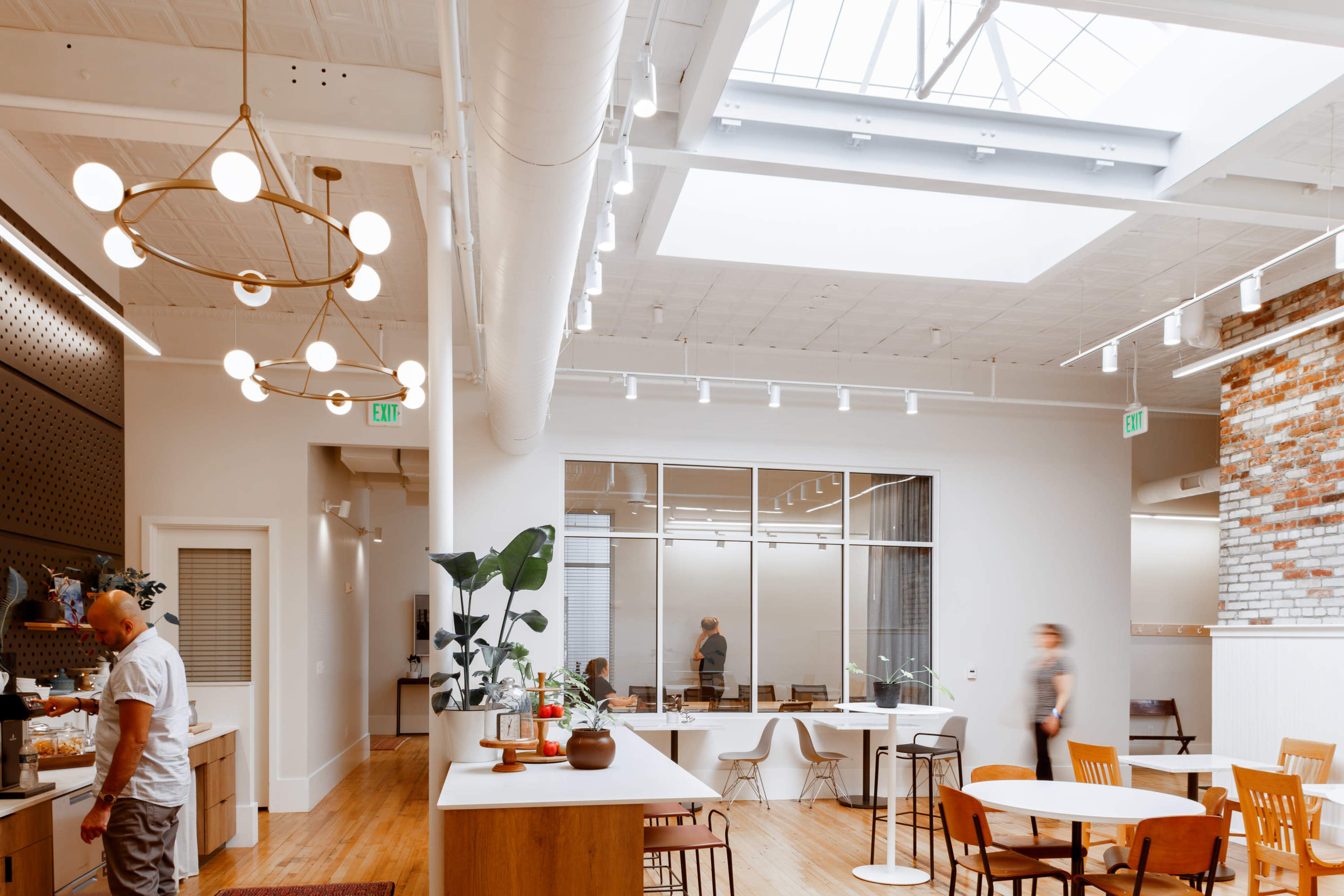 A spacious, modern office features a kitchen area, large windows, and a mix of furniture, with people casually interacting.