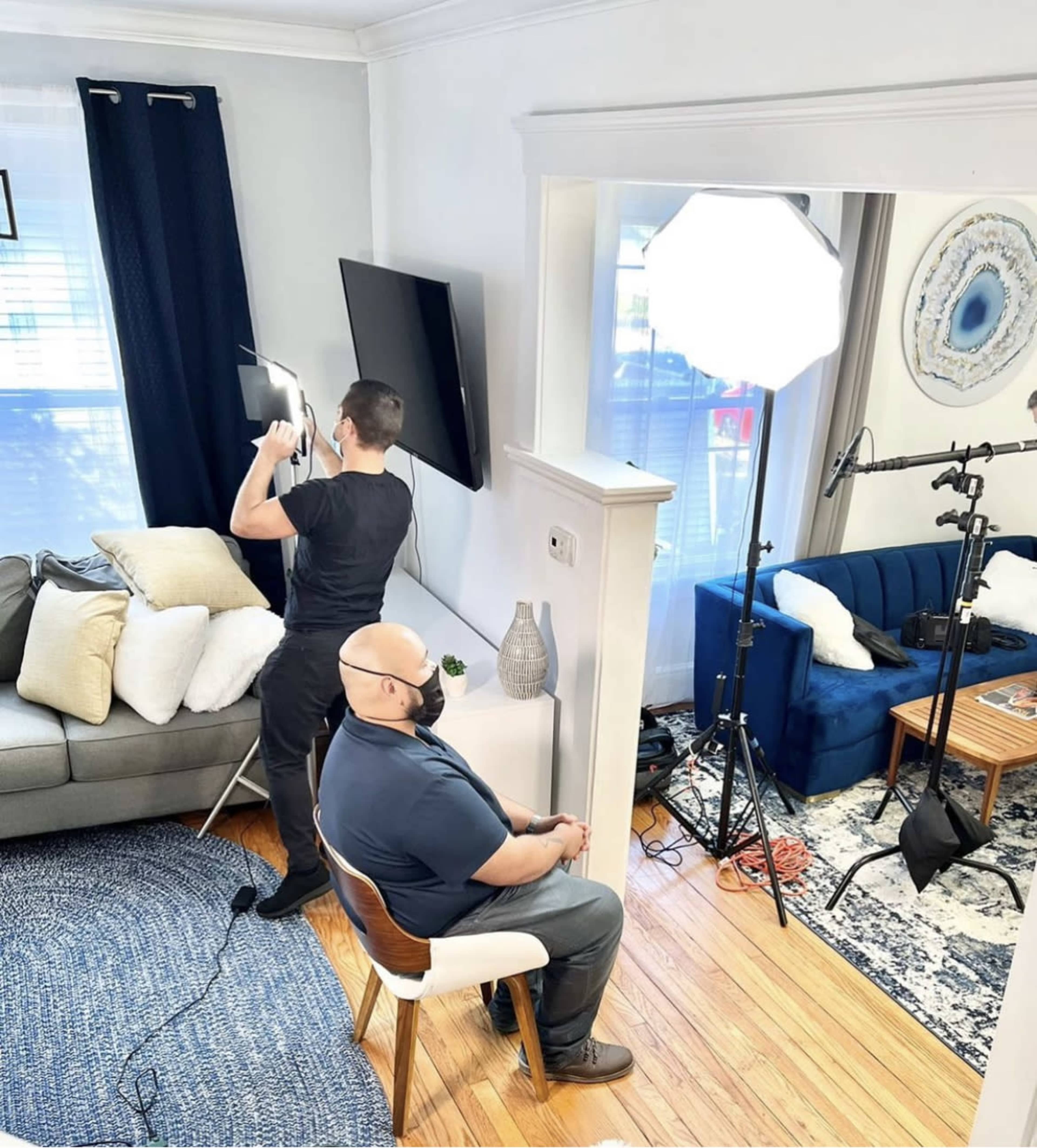 A man is sitting on a chair in a living room while another man adjusts a lighting setup nearby.