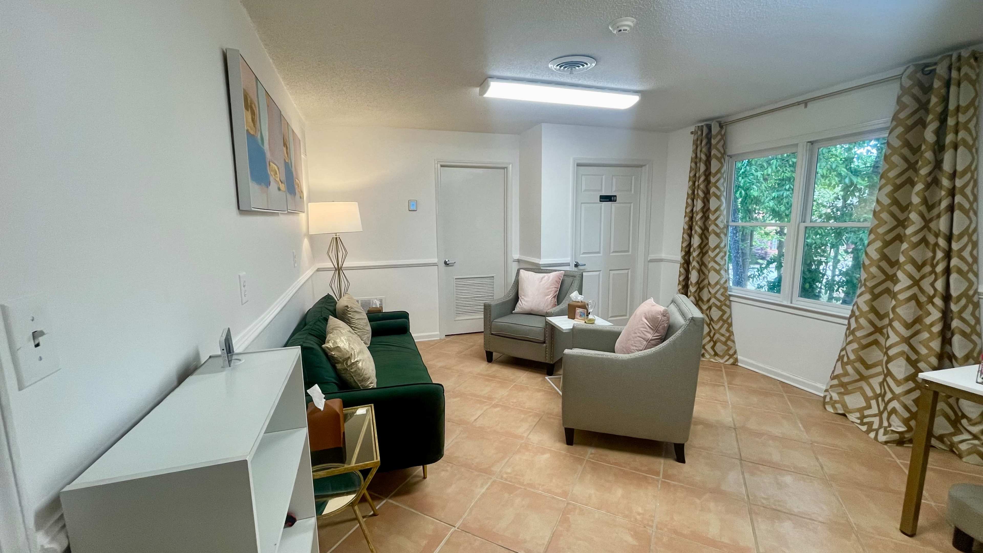 A small living area featuring a green sofa, two gray armchairs, a lamp, and a side table, with curtains and windows allowing natural light.