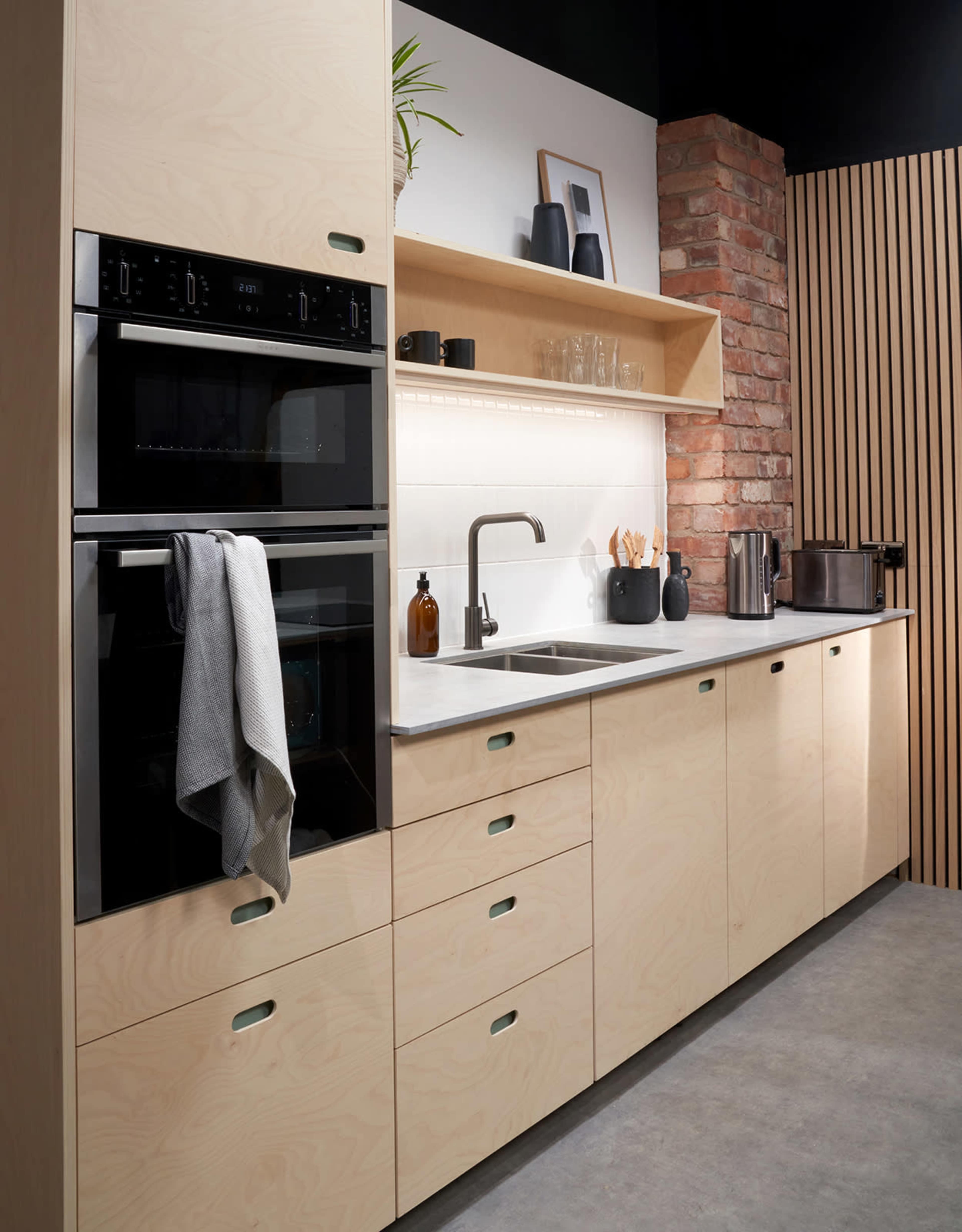 A modern kitchen featuring beige cabinetry, built-in black ovens, a minimalist sink, and a wooden accent wall.