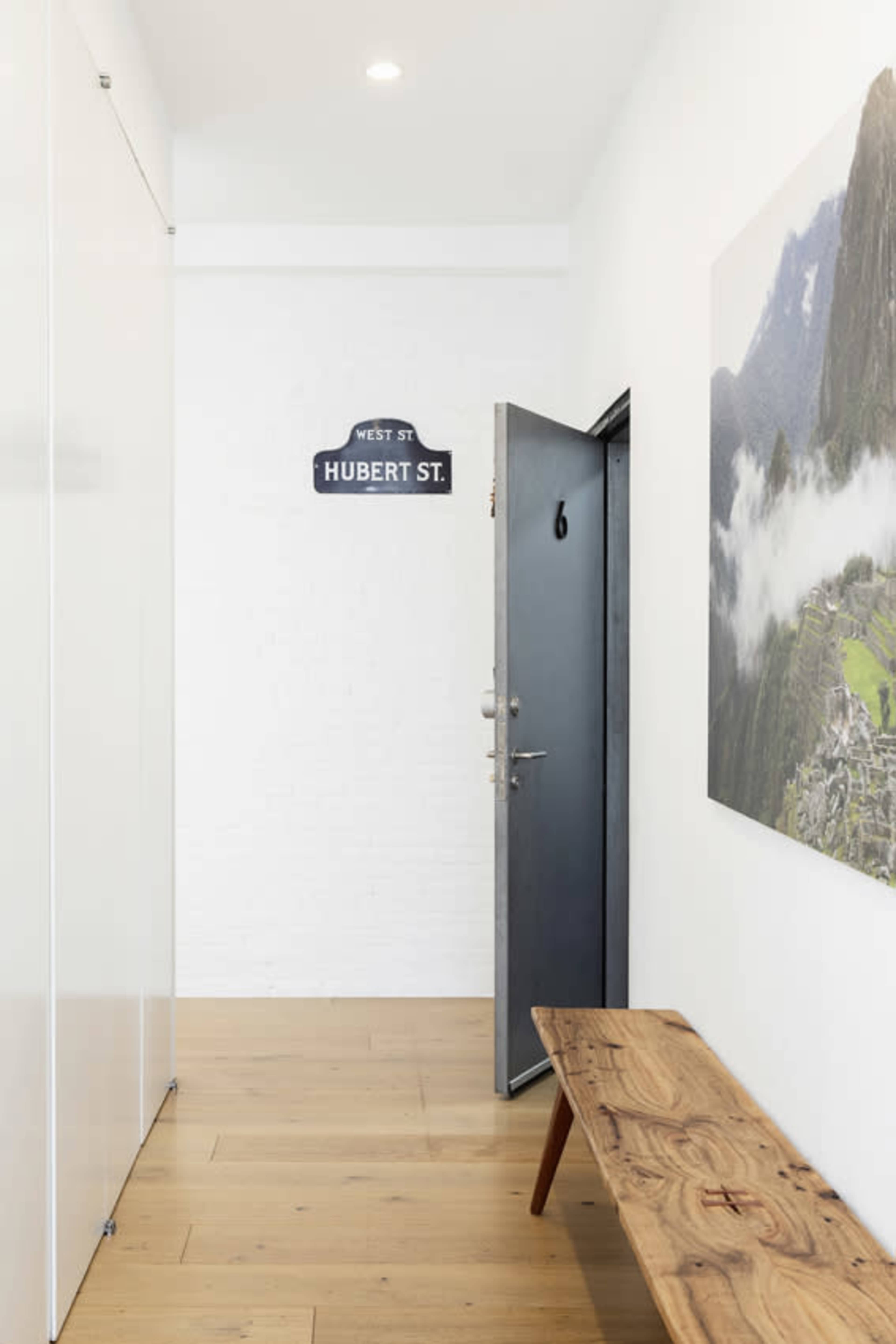 A minimalist hallway with a dark door labeled "6" and a wooden bench against a white wall, featuring a landscape photograph and a street sign that reads "WEST ST. HUBERT ST."