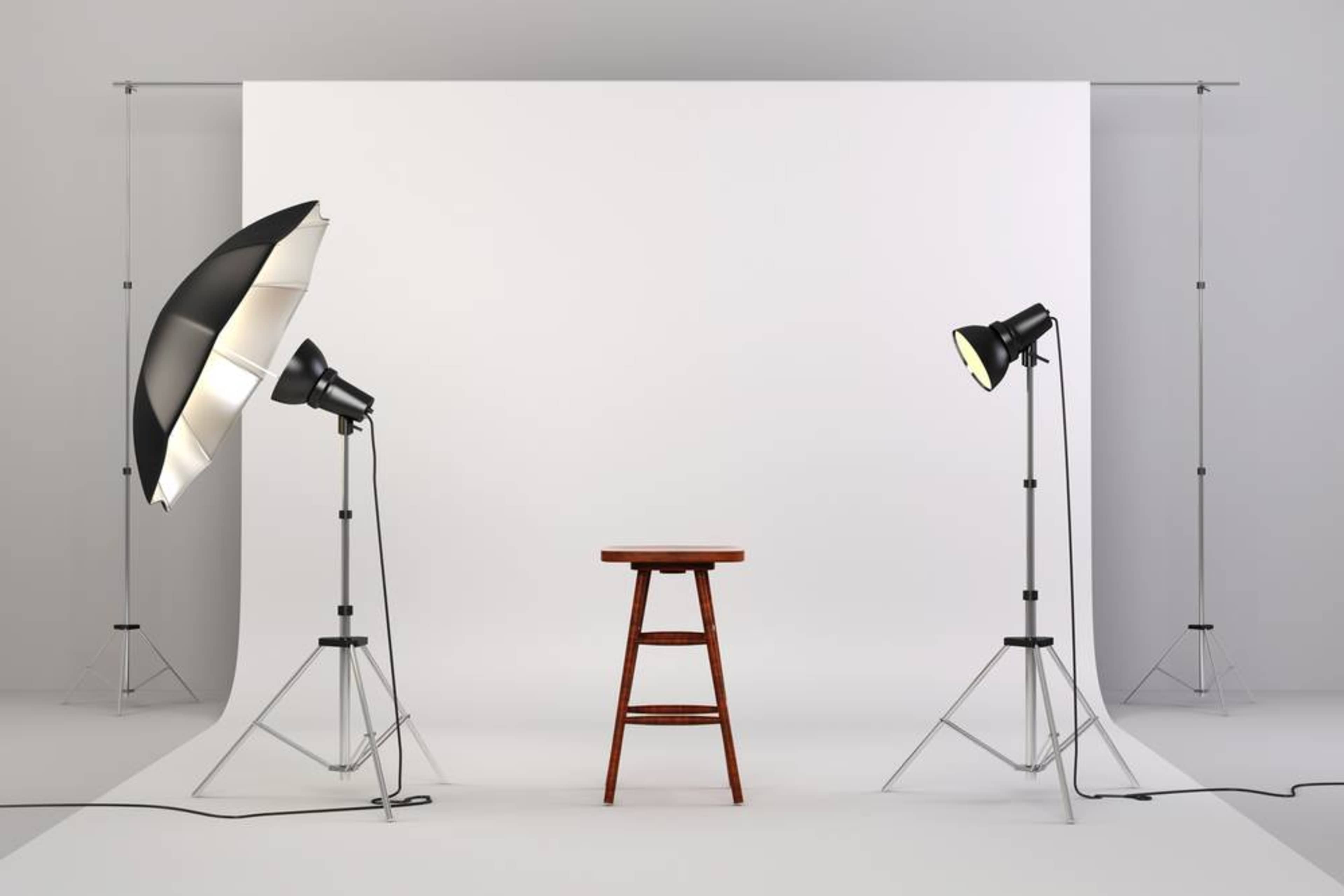 A photography studio setup featuring a wooden stool placed in front of a white backdrop, illuminated by two lighting fixtures on adjustable stands.