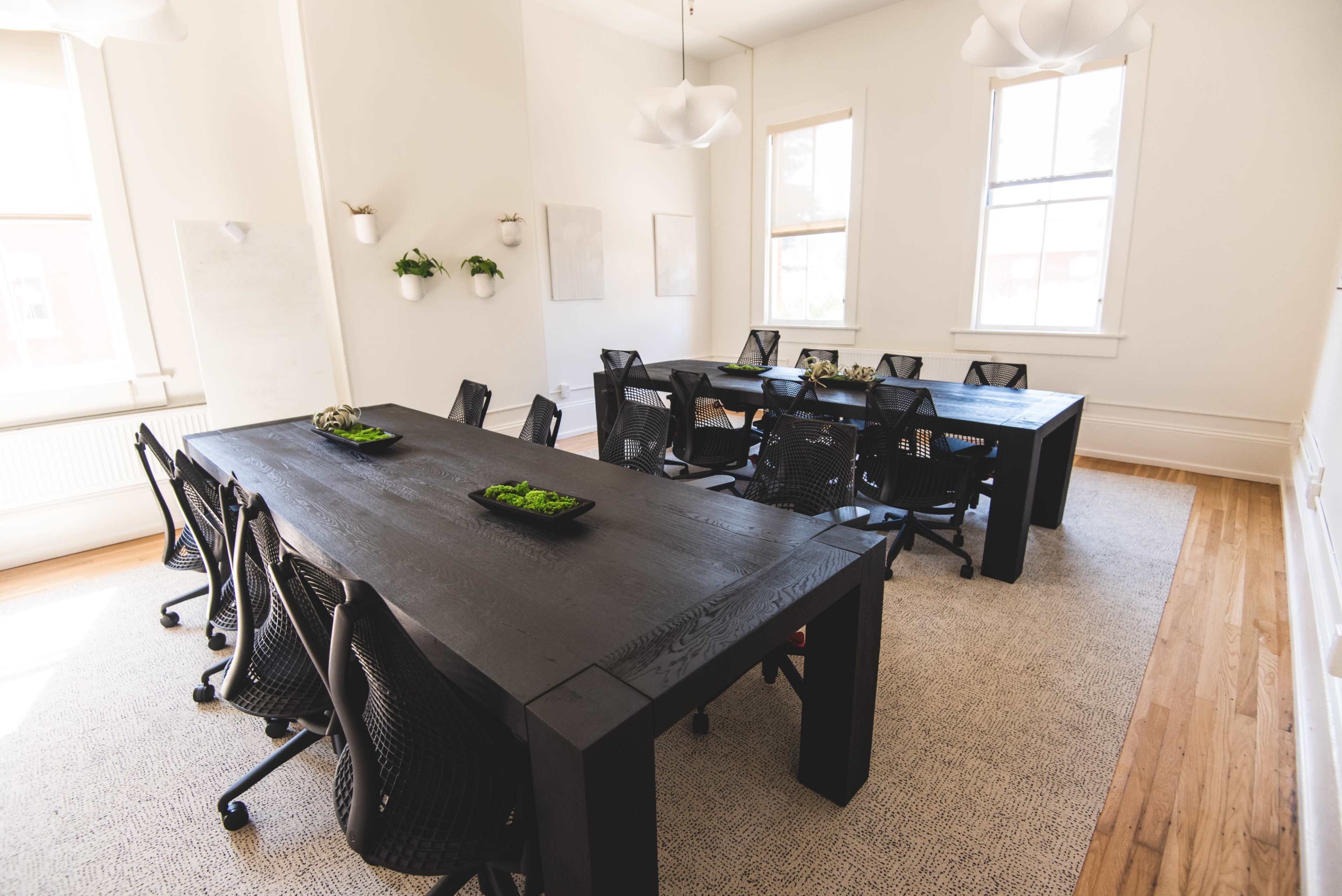 The image shows a spacious meeting room with two large black tables surrounded by black office chairs, featuring bright windows and minimalist decor.