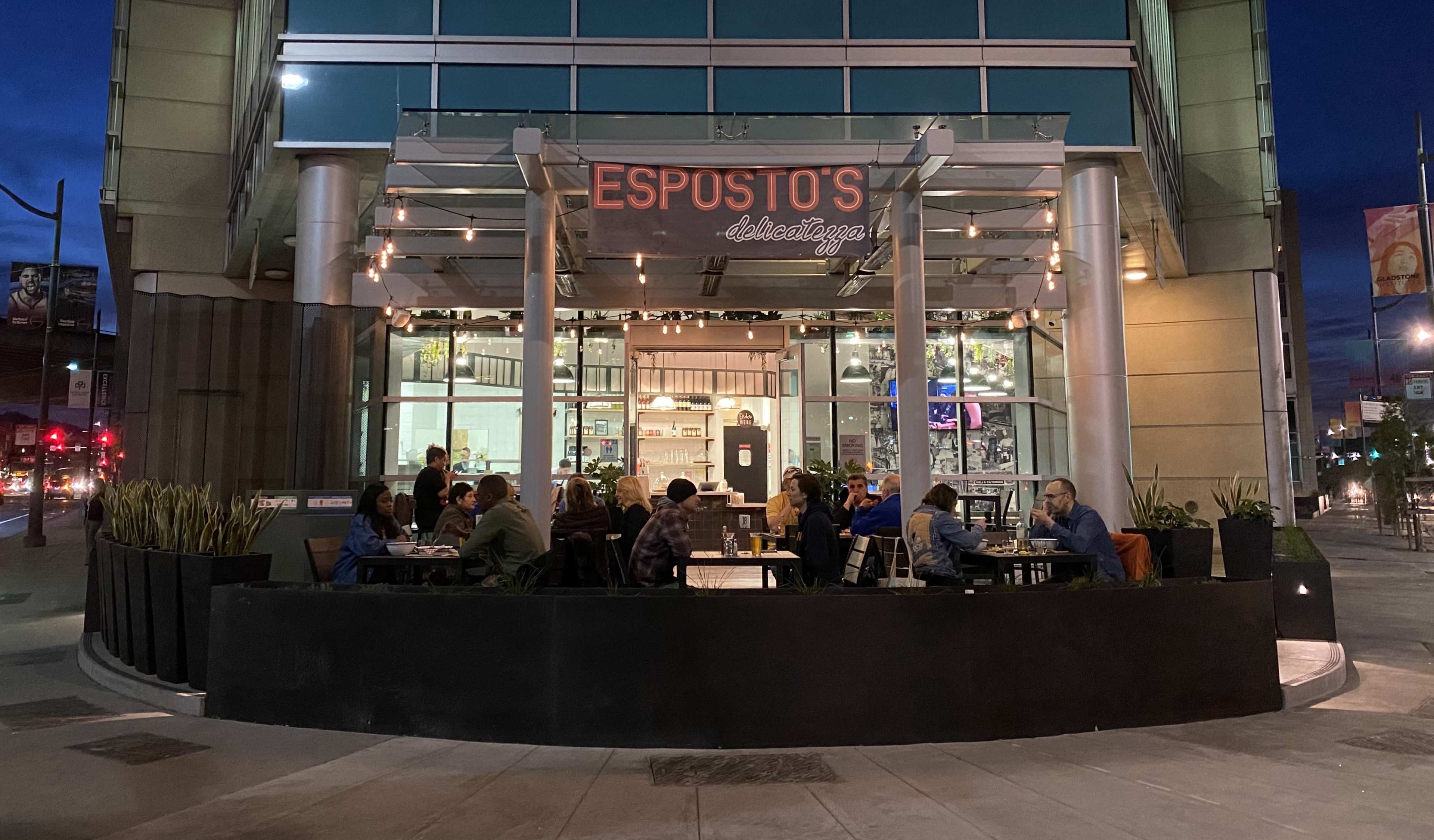 A restaurant patio is filled with patrons dining at tables, while the building's facade is illuminated during dusk.