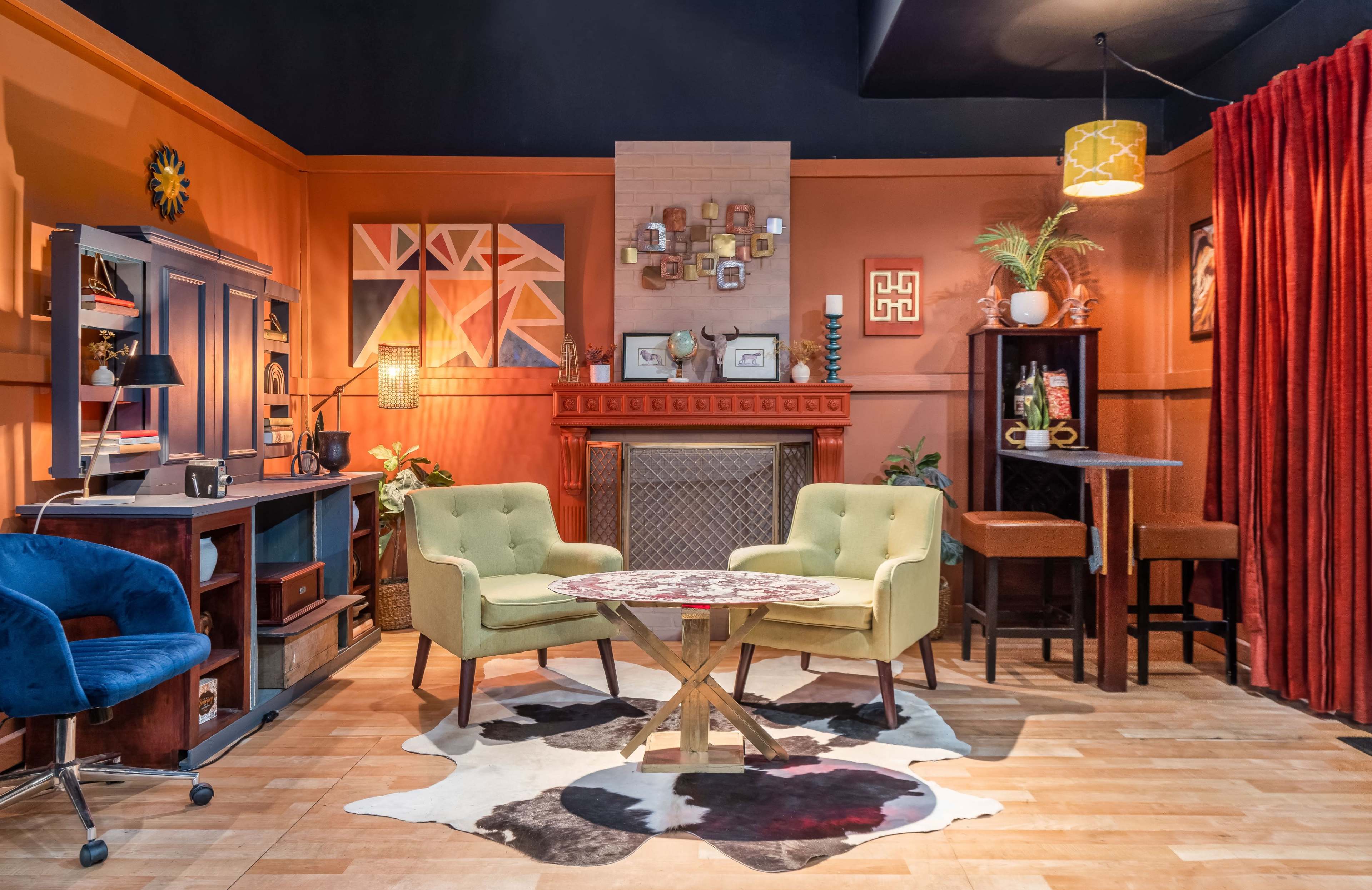 The scene features a cozy mid-century modern living room with a fireplace, two armchairs, a small round table, a bar area, and various decorative elements on the walls.