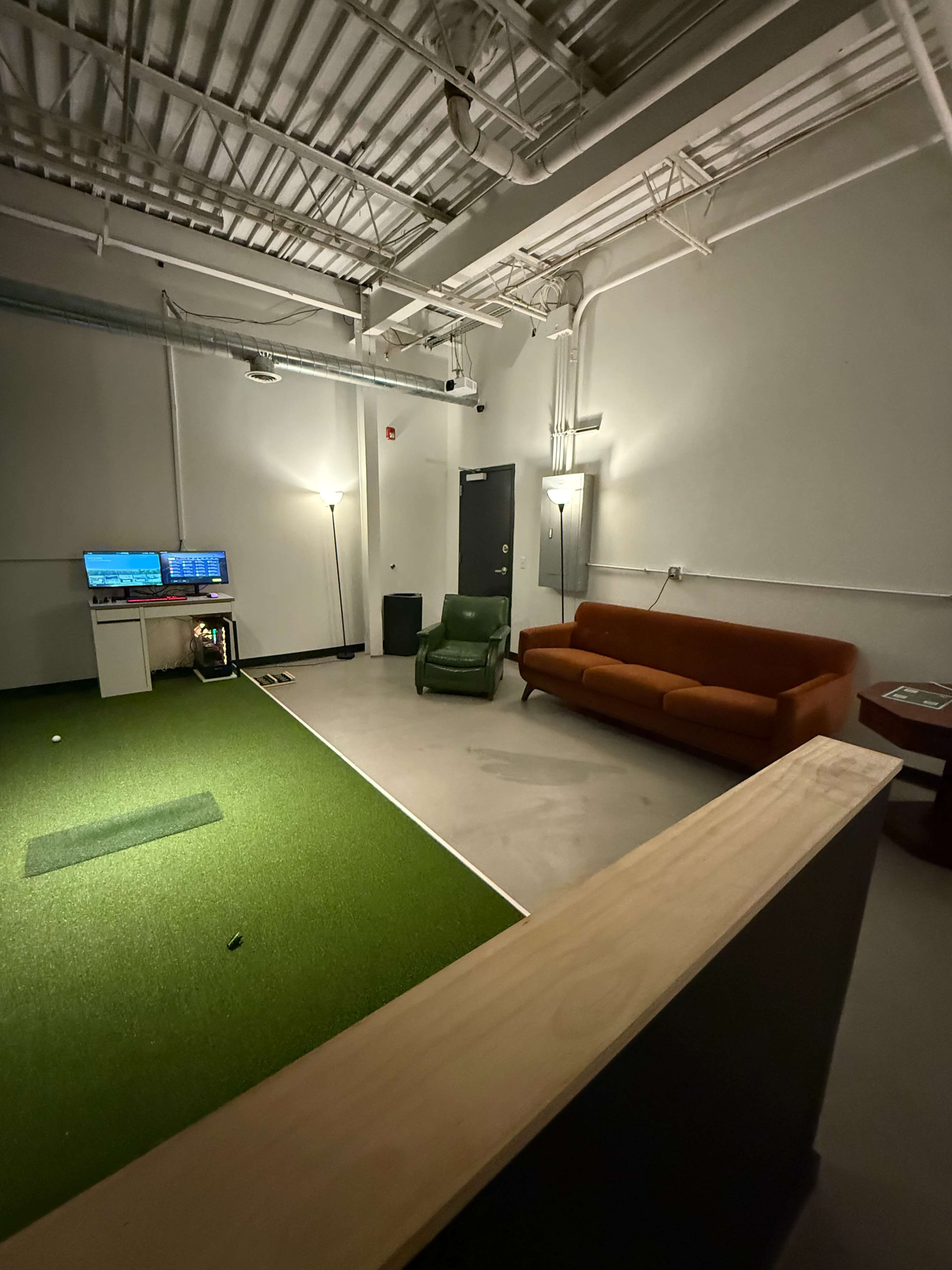 The image shows a dimly lit room with a green turf area, a couch, a chair, and a television displaying a golf-related screen.