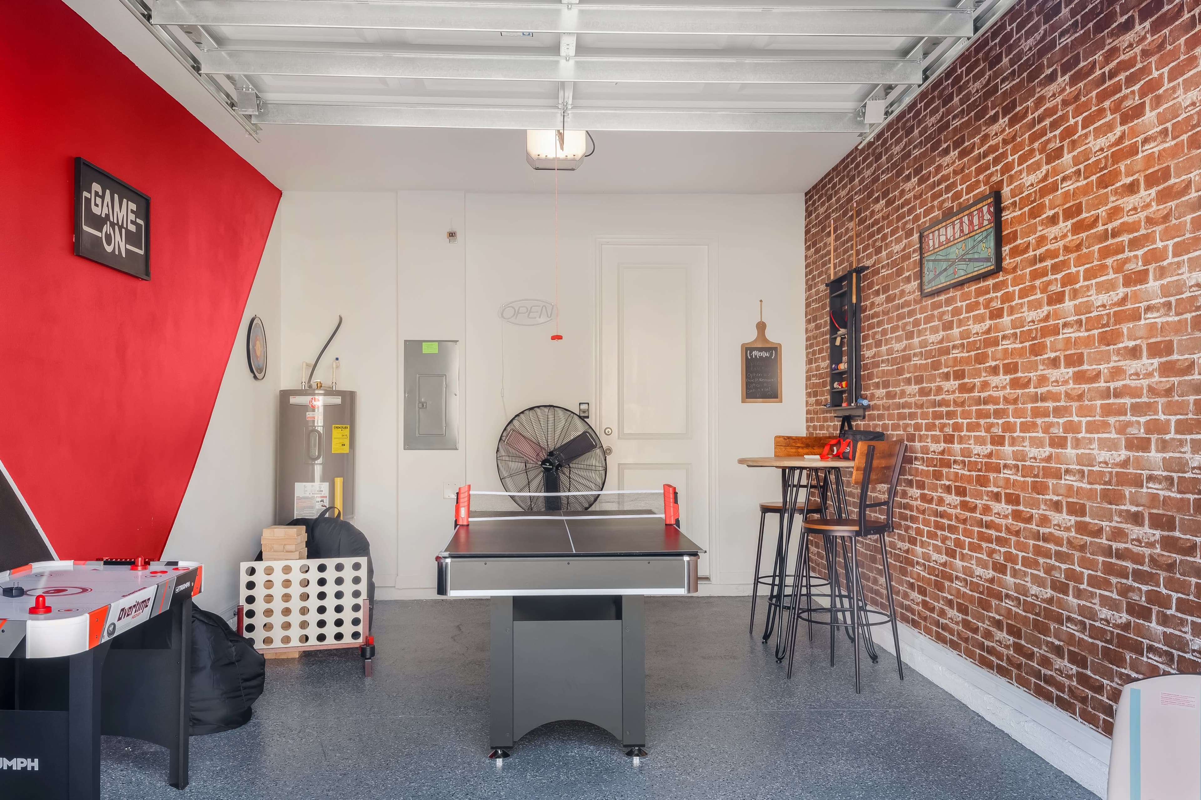 The image shows a garage game room featuring an air hockey table, a bar-height table with stools, exposed brick walls, and a red accent wall.