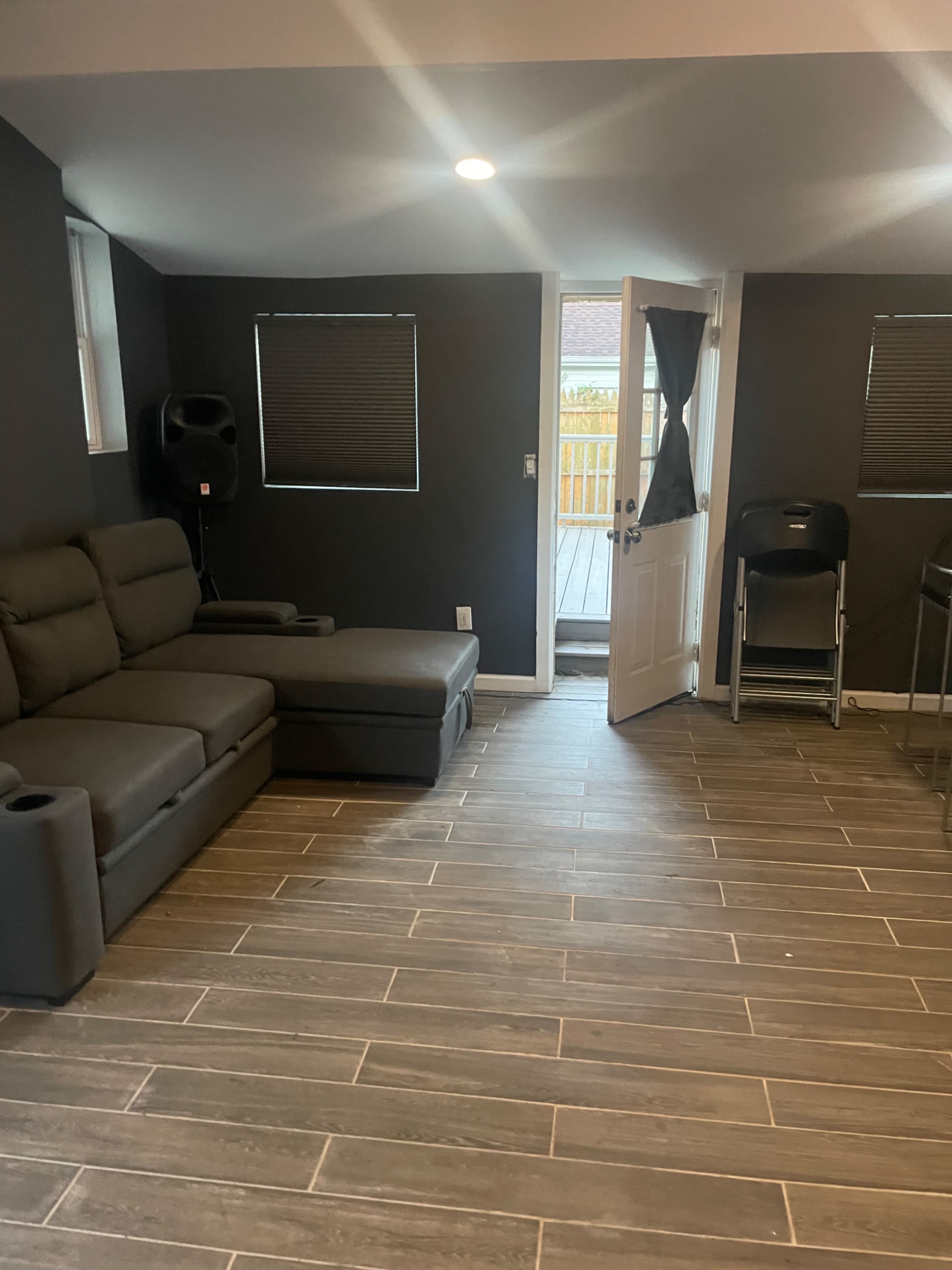 A living room with a gray sectional sofa, a door leading to a deck, and a set of stacked folding chairs against the wall.