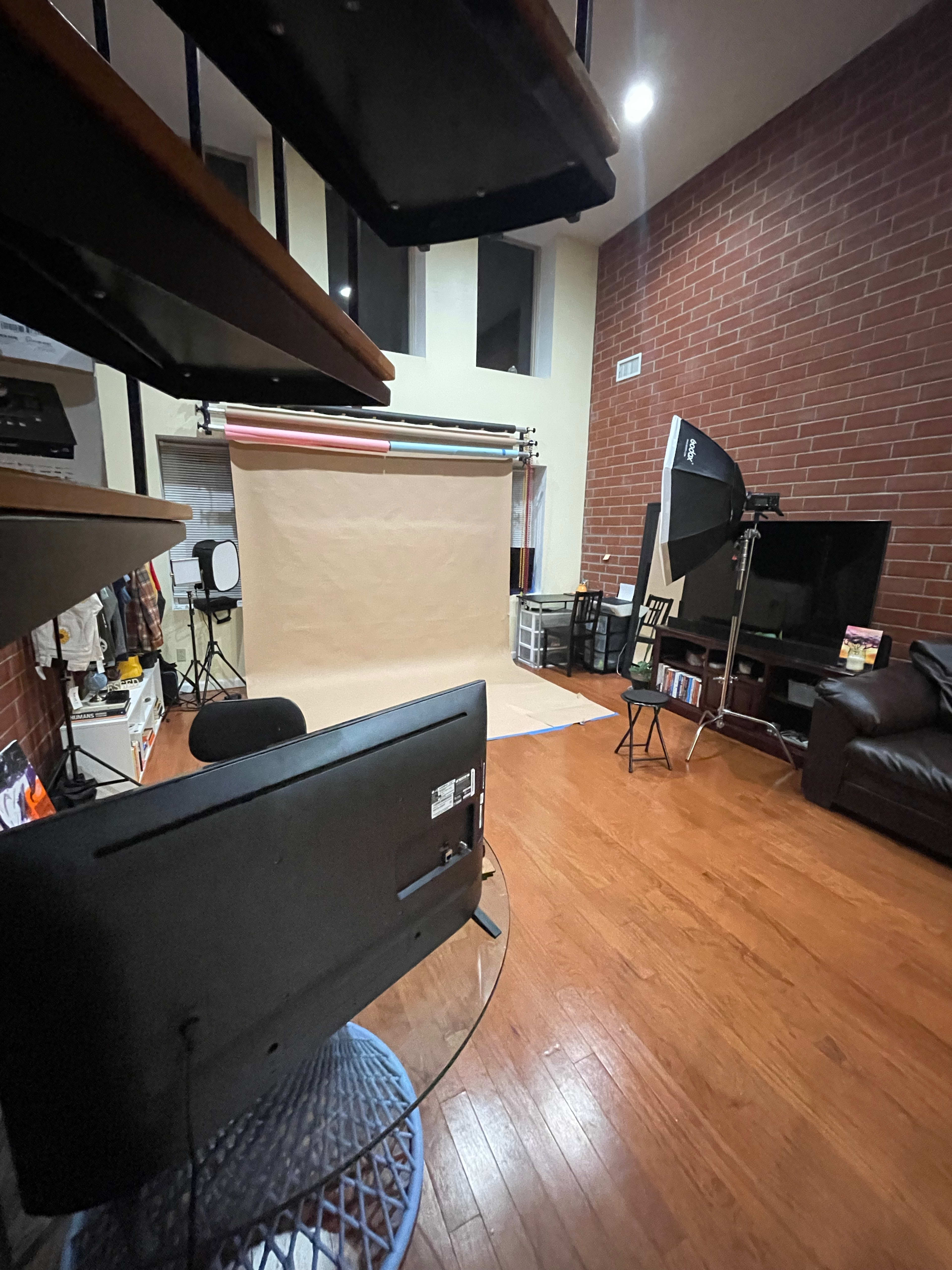 An interior studio setting with a seamless backdrop, lighting equipment, and a living area featuring a television and couch.