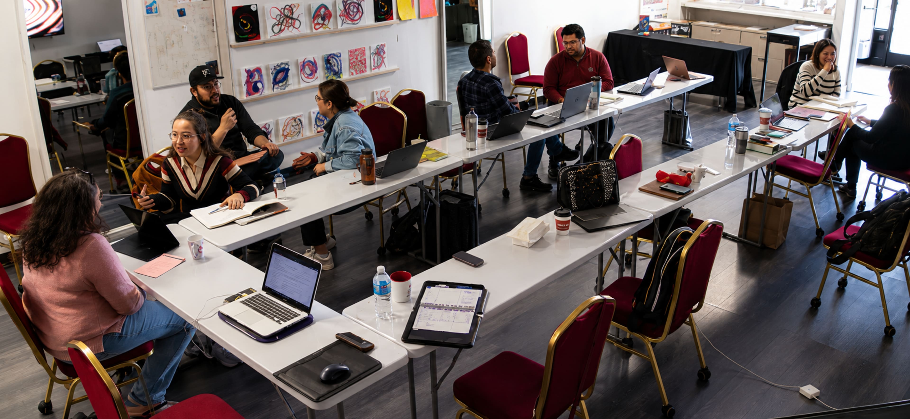A group of people engages in discussions and work at a long table in a brightly lit room with various laptops and documents spread out.