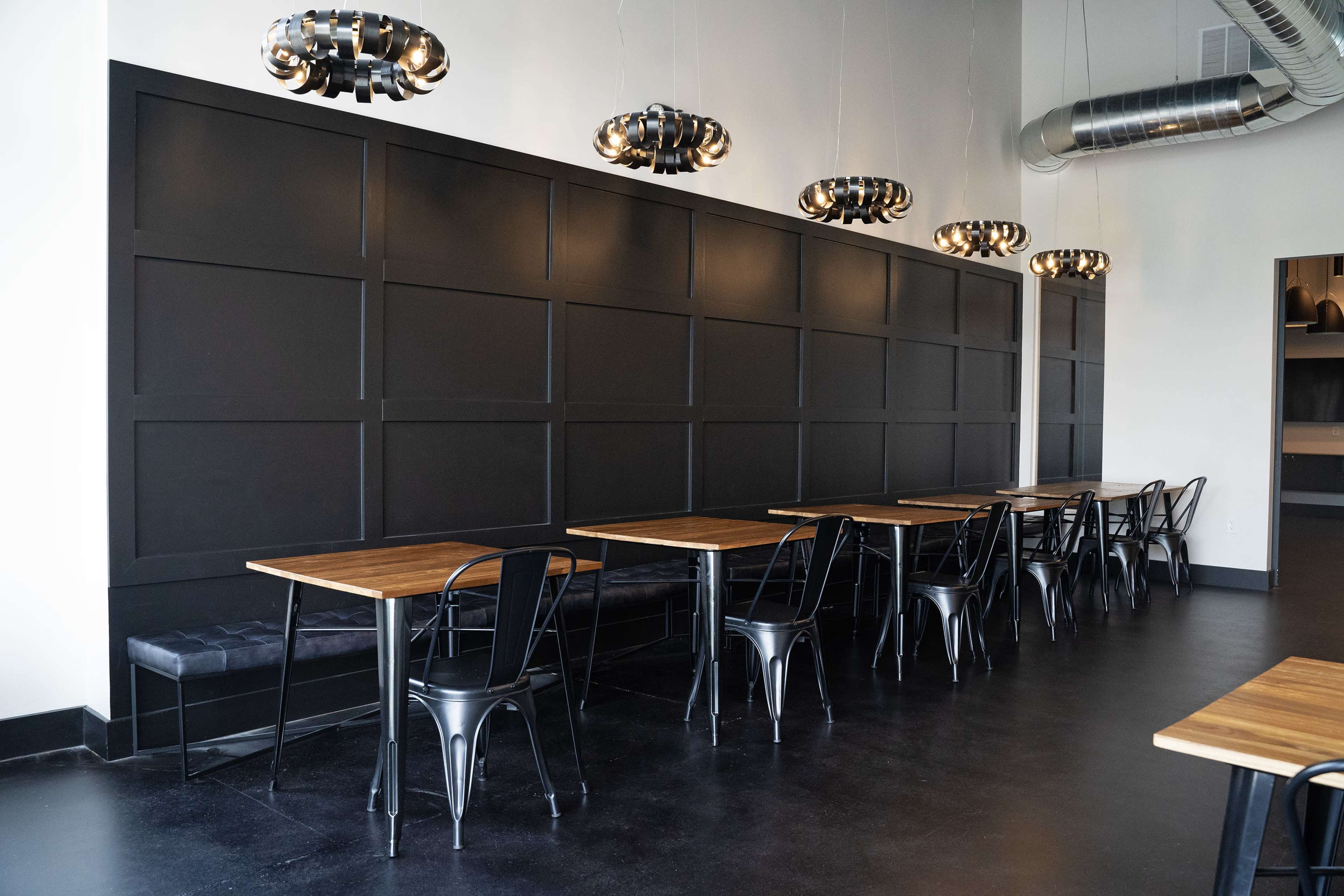 A modern, minimalist restaurant interior with black walls, wooden tables, and hanging light fixtures.