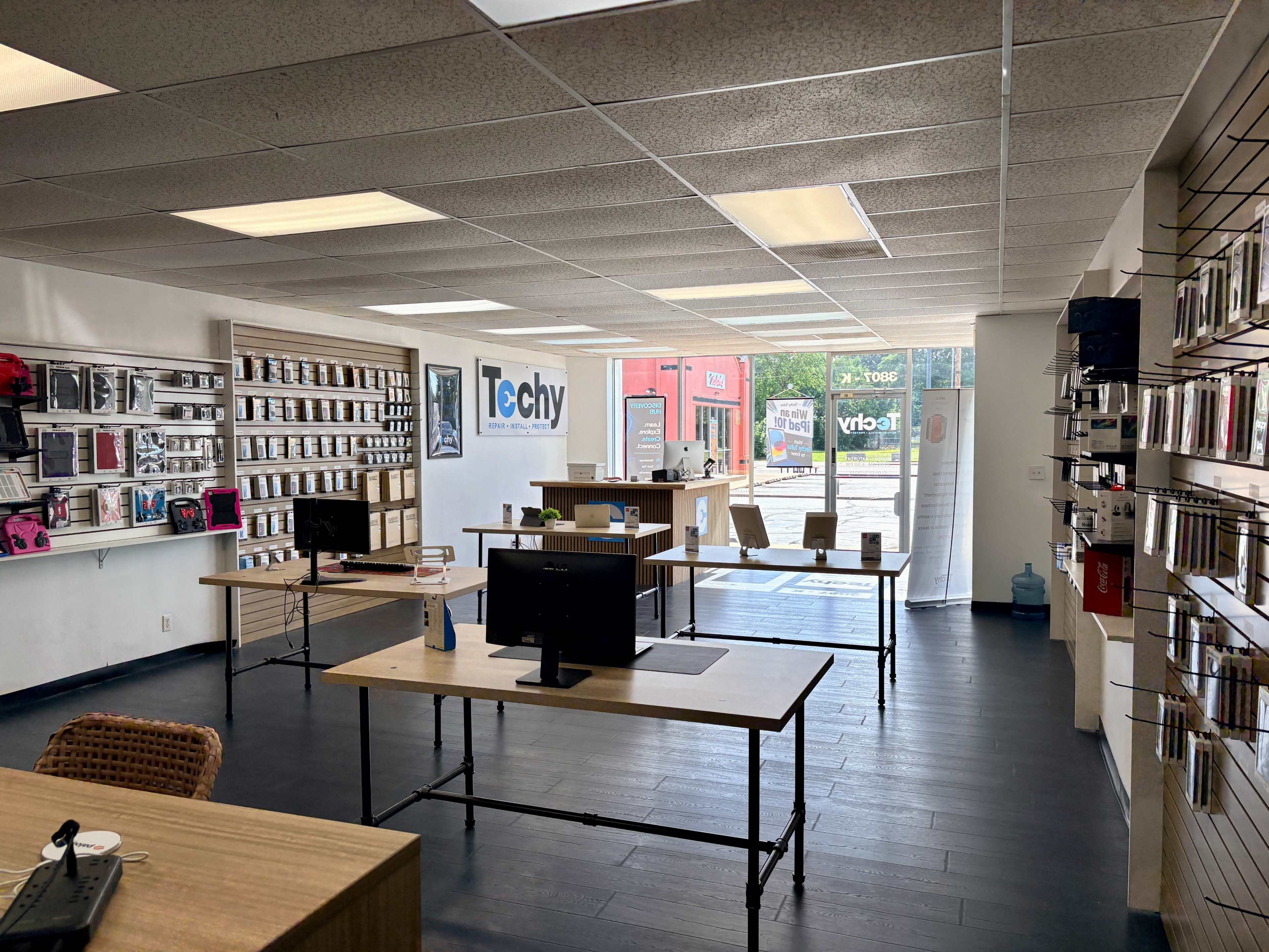 The image shows a technology retail store with wooden tables displaying products, wall shelves filled with merchandise, and large windows providing natural light.