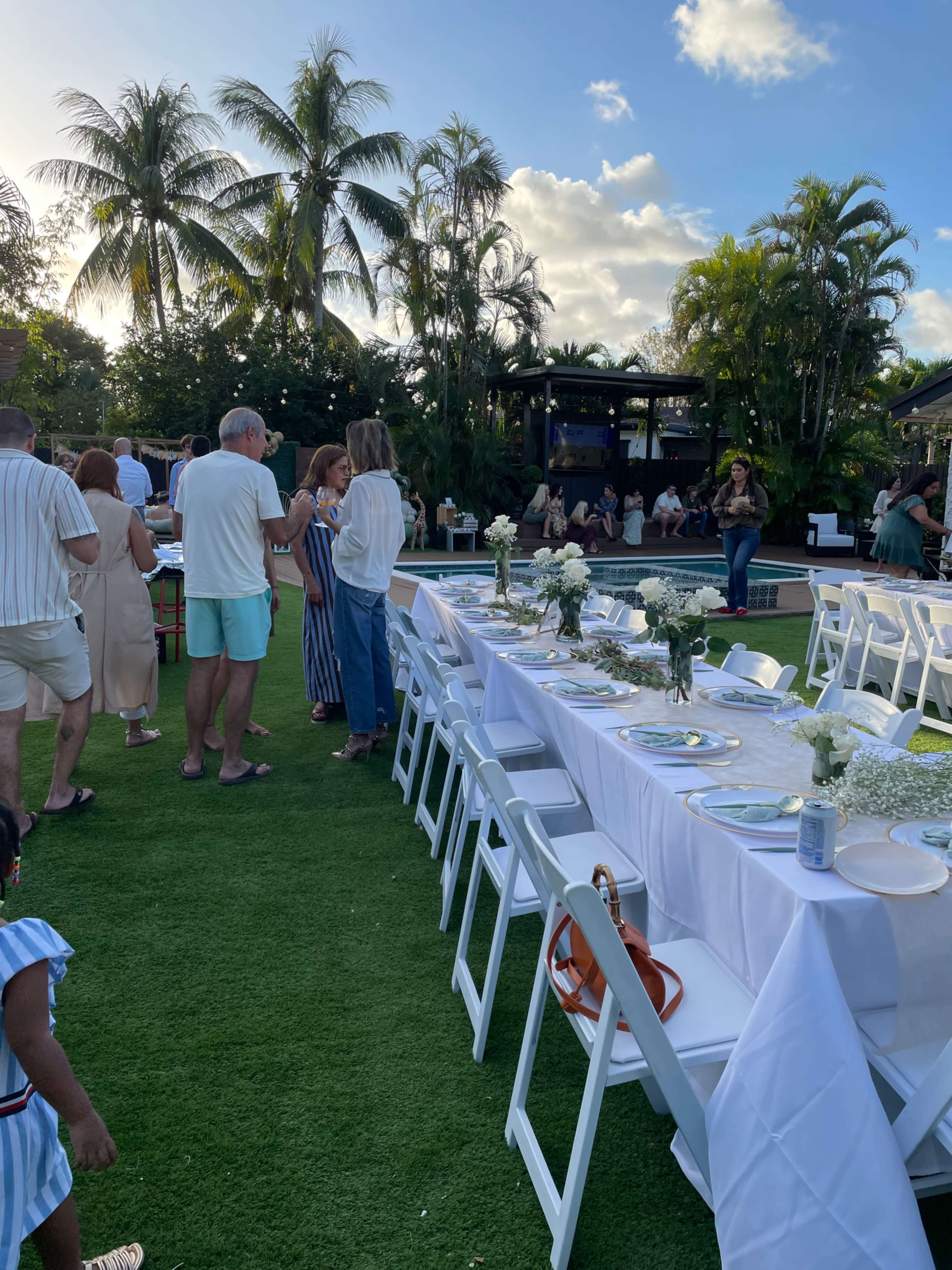 A long table is set up outdoors with white tableware and floral centerpieces, surrounded by people mingling in a tropical garden setting.