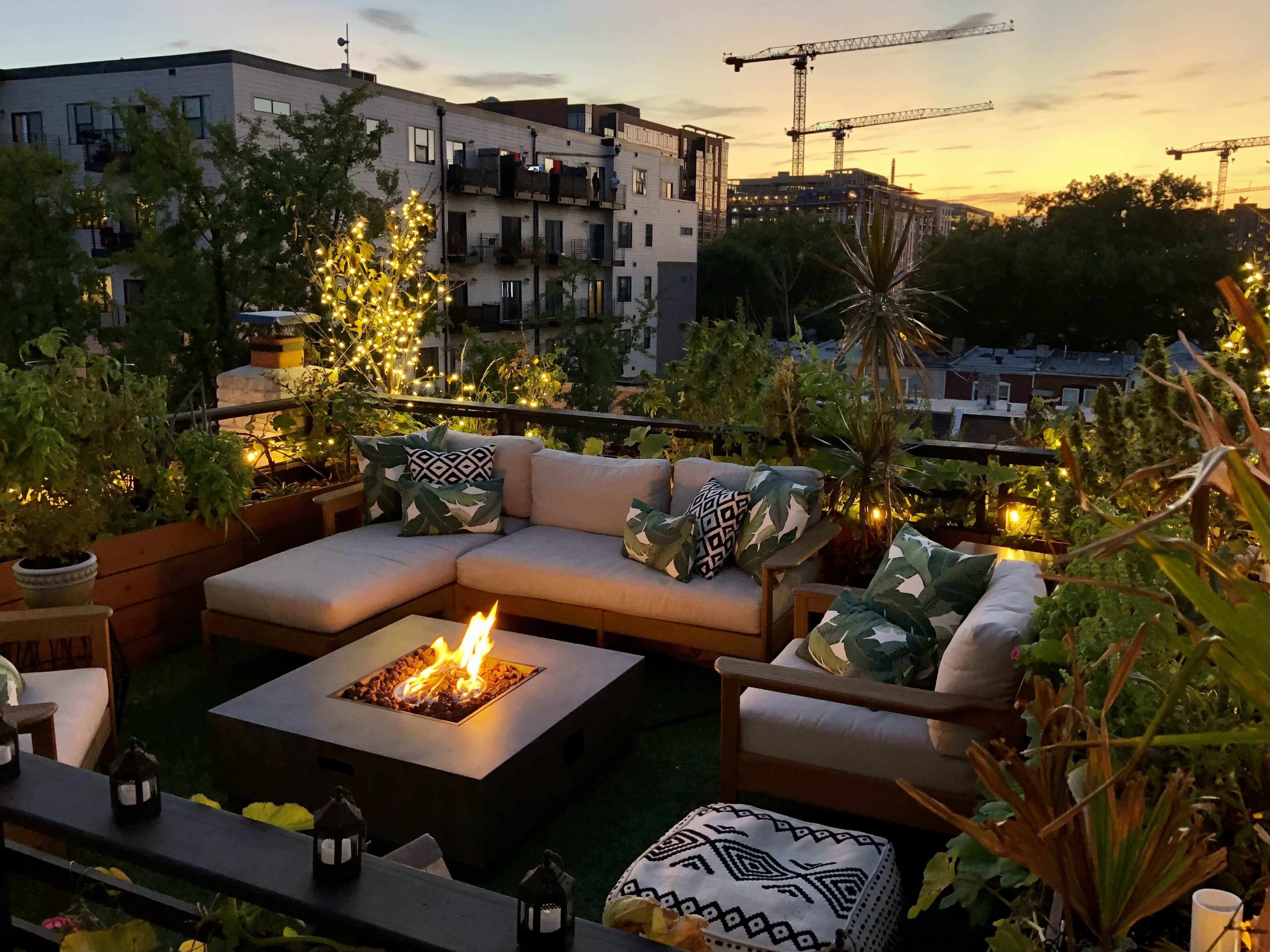 A rooftop terrace features a seating area with a fire pit, surrounded by greenery and decorative lights, against a sunset backdrop with construction cranes in the distance.