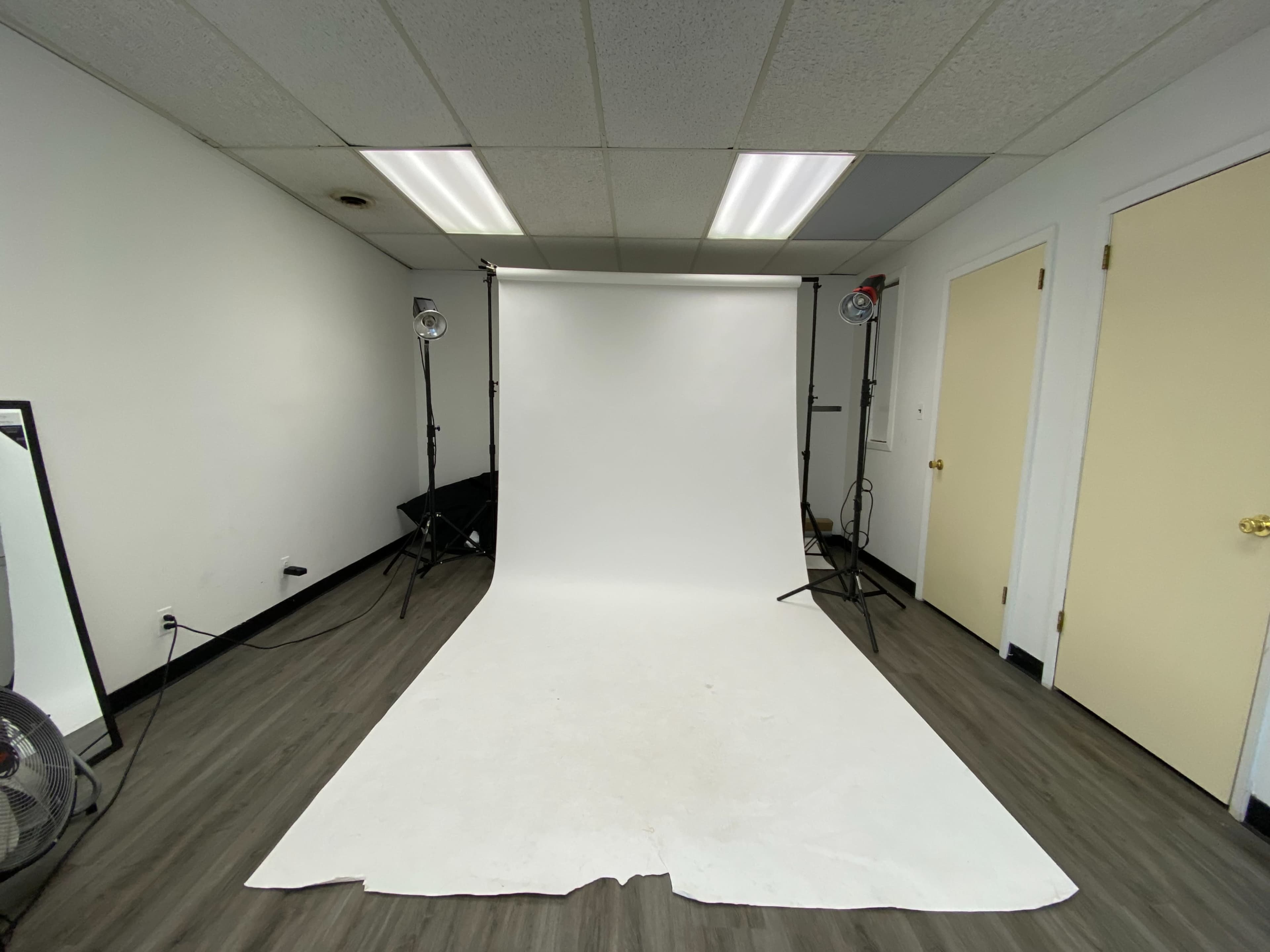 The image shows a photo studio with a white backdrop, two lights on stands, and a fan in the corner.