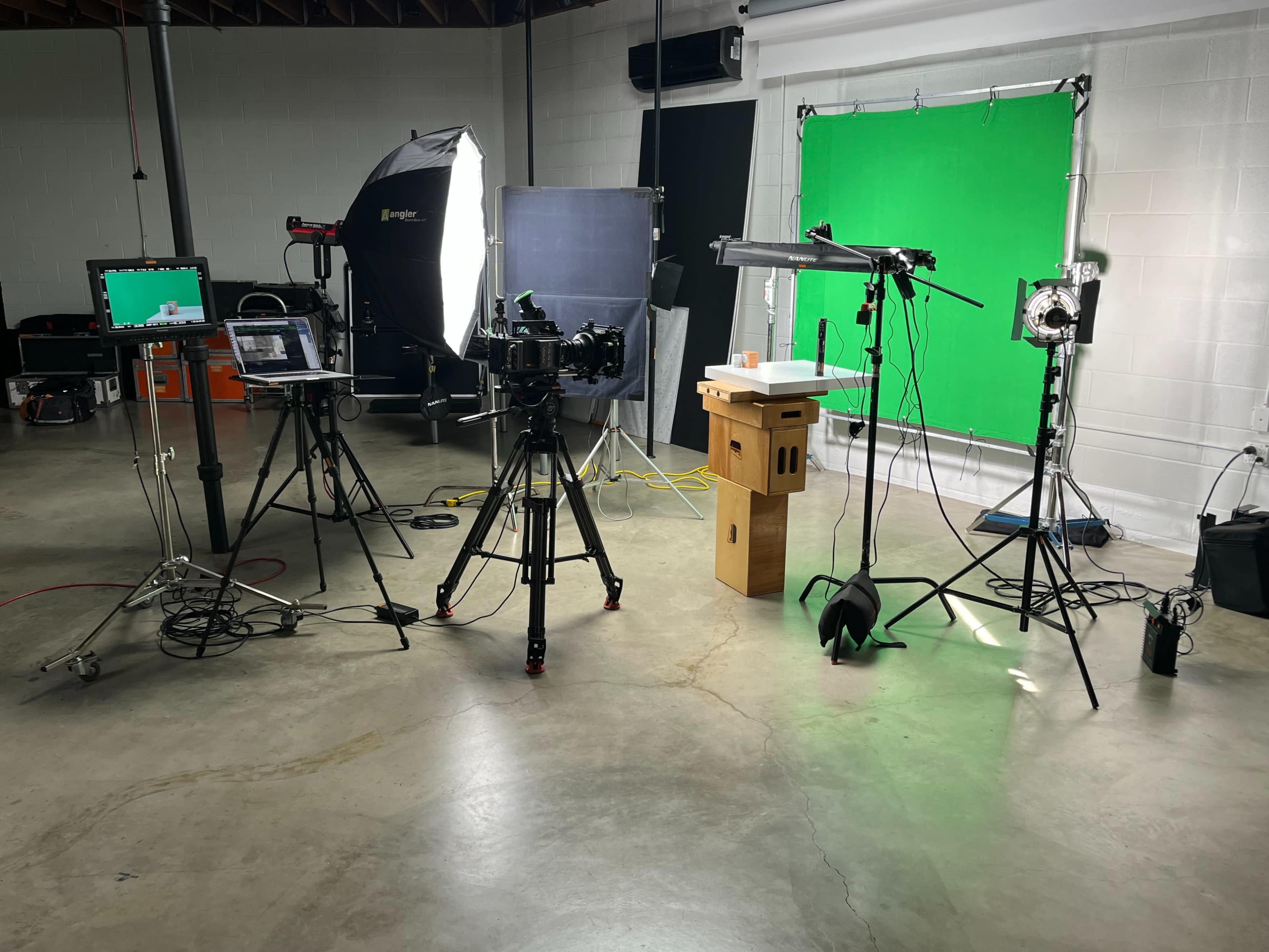The image shows a video production studio setup with various cameras, lighting equipment, and a green screen in the background.