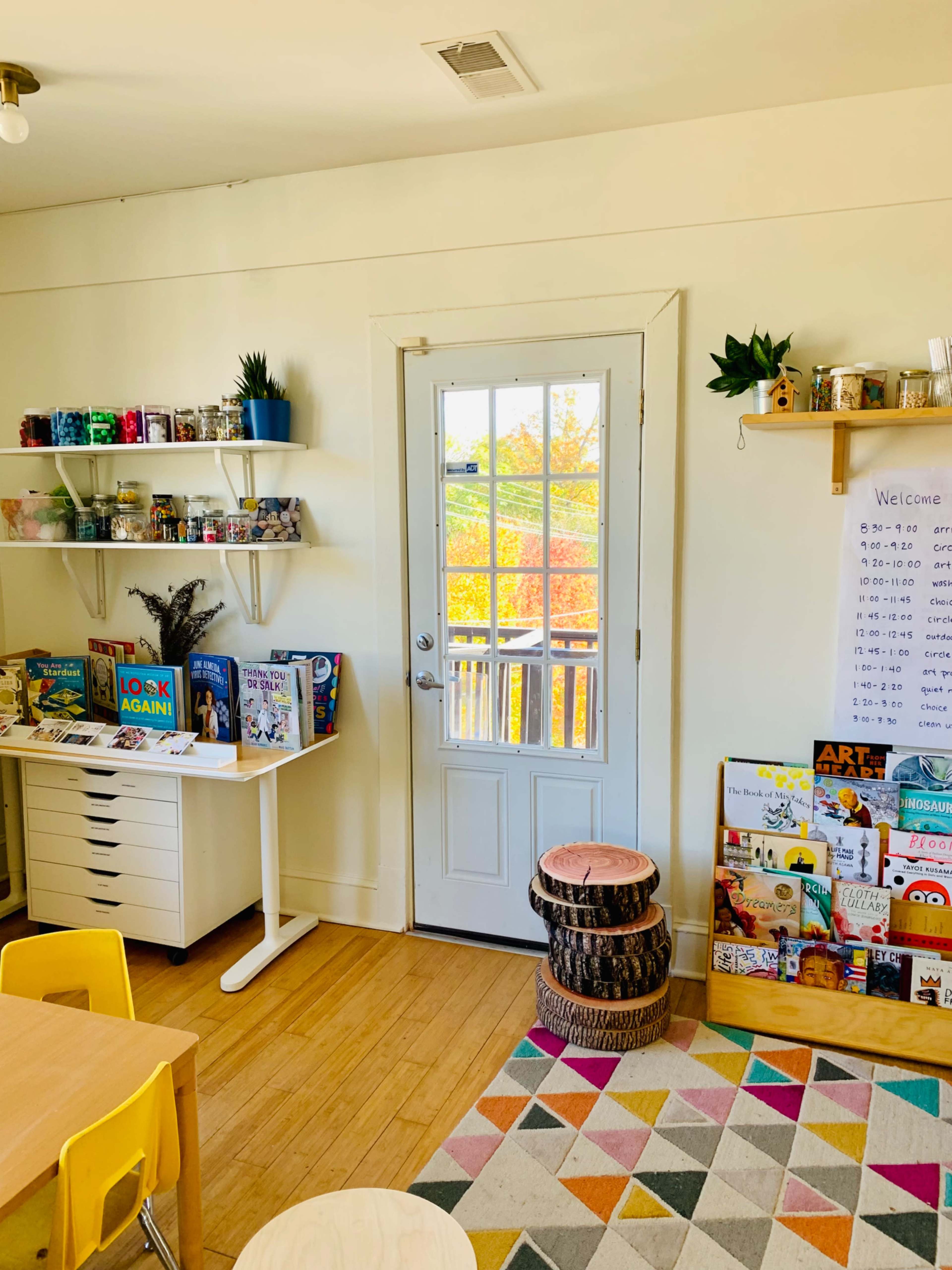 A brightly lit room features a white door leading to an outdoor view, bookshelves stocked with colorful books and materials, and a patterned rug under a table and chairs.