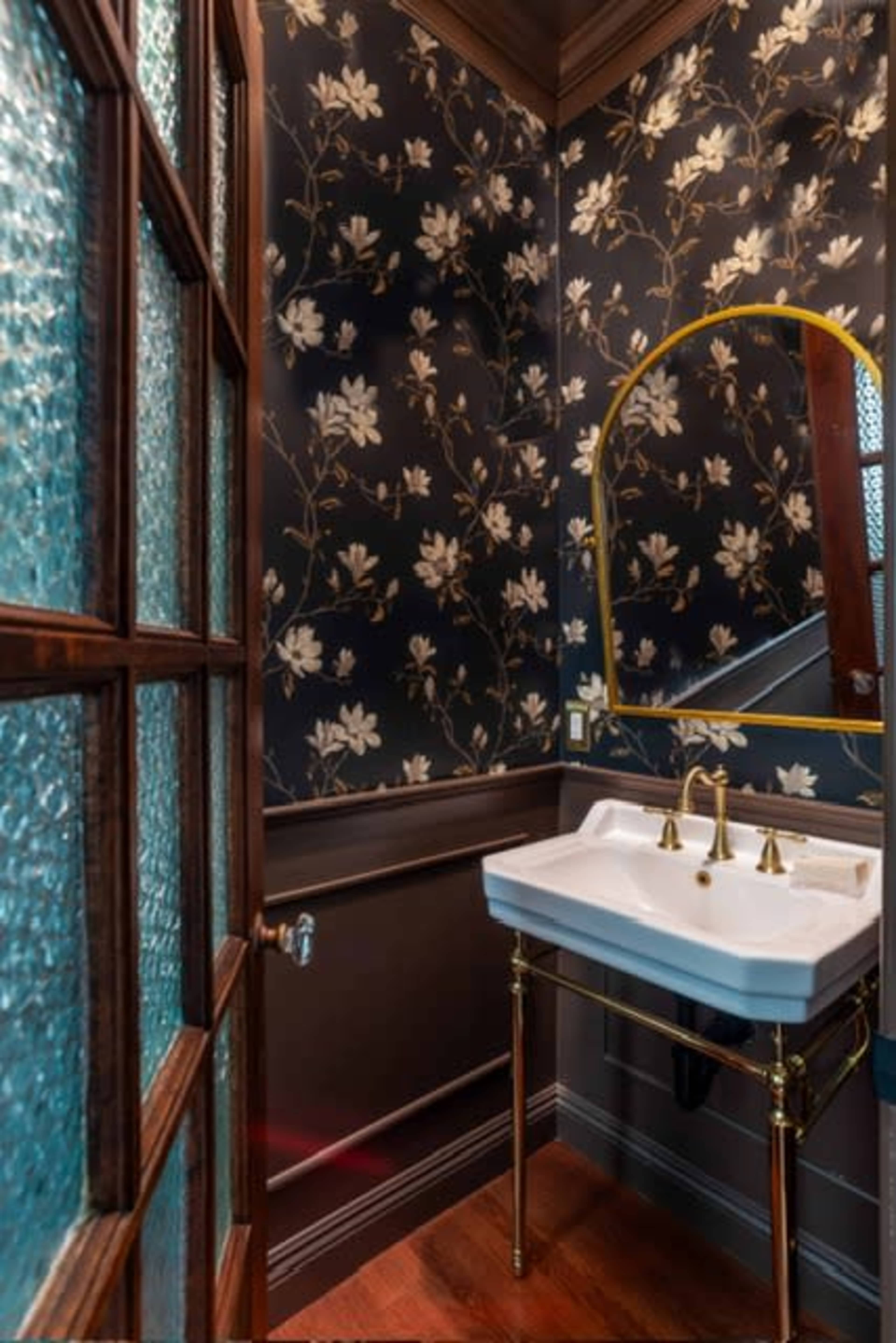 A small bathroom features a floral-patterned black and gold wallpaper, a white sink with gold faucets, and a mirror with an arched frame.