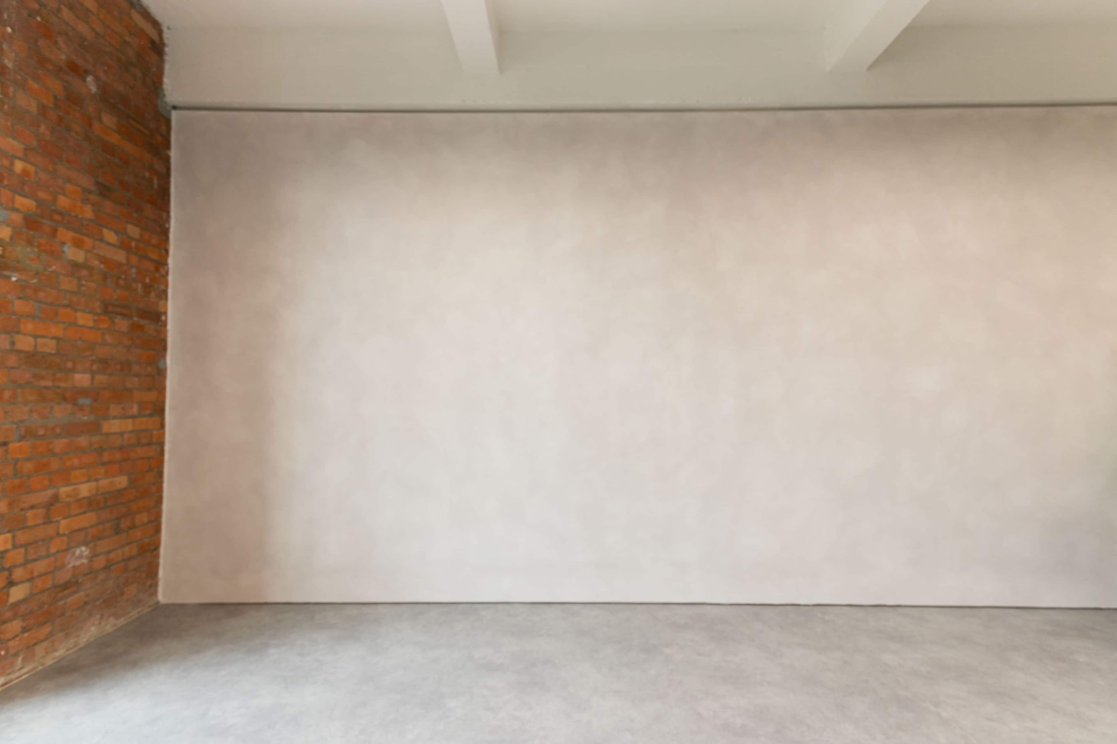 The image shows an empty room with a brick wall on one side and a smooth, light-colored wall on the opposite side.