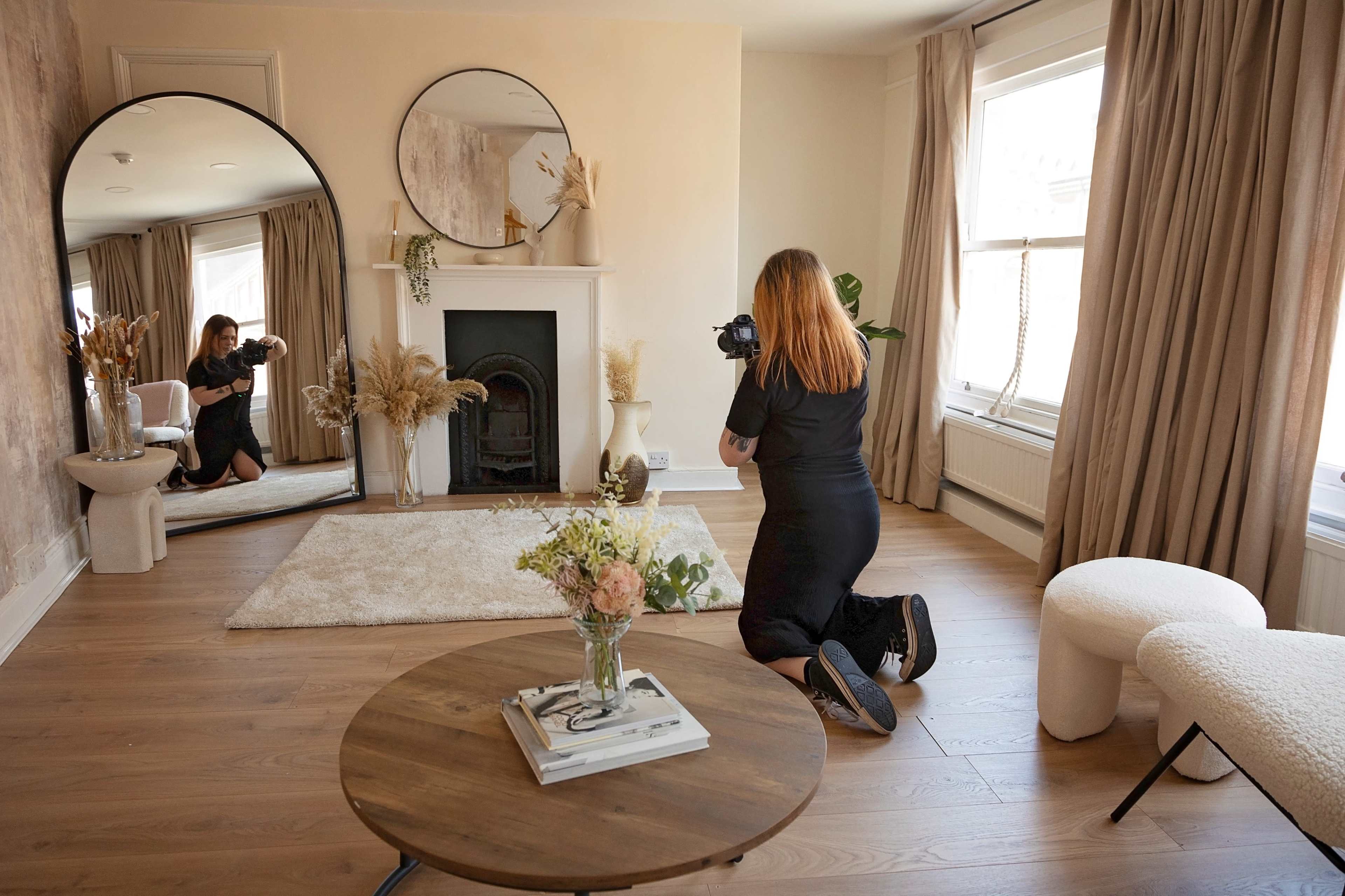 A woman is kneeling on the floor while taking photographs of a stylish living room featuring a fireplace, large mirrors, and decorative plants.