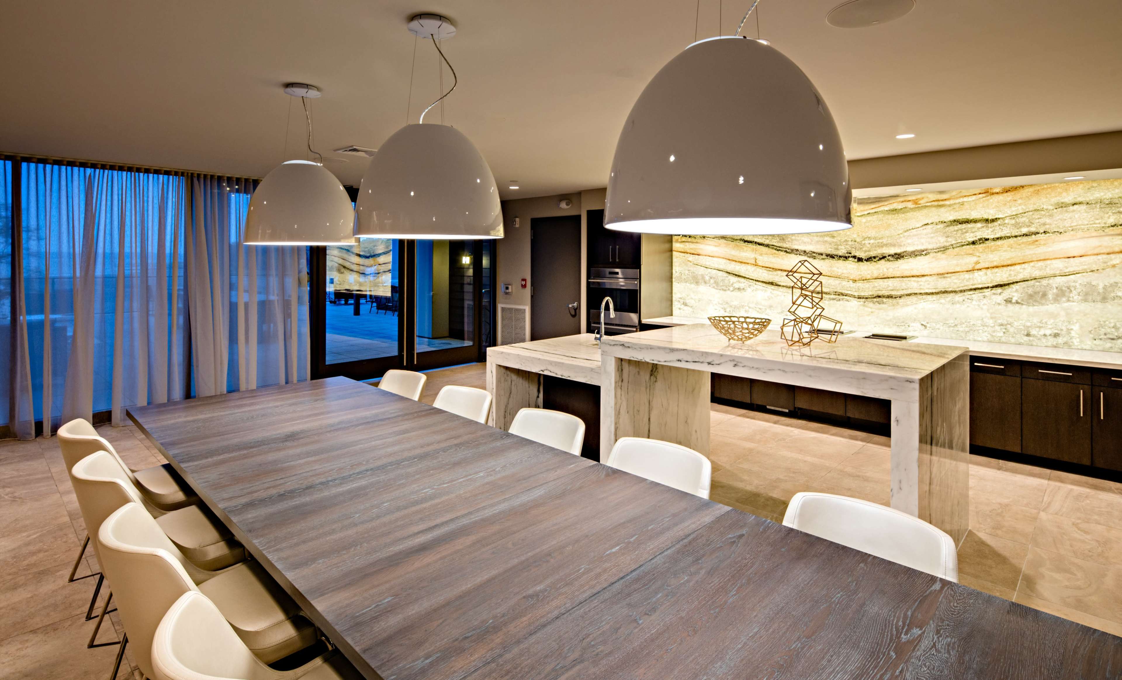 A modern kitchen features a large wooden dining table surrounded by white chairs, with pendant lights above and a stone countertop illuminated by a backlit wall.