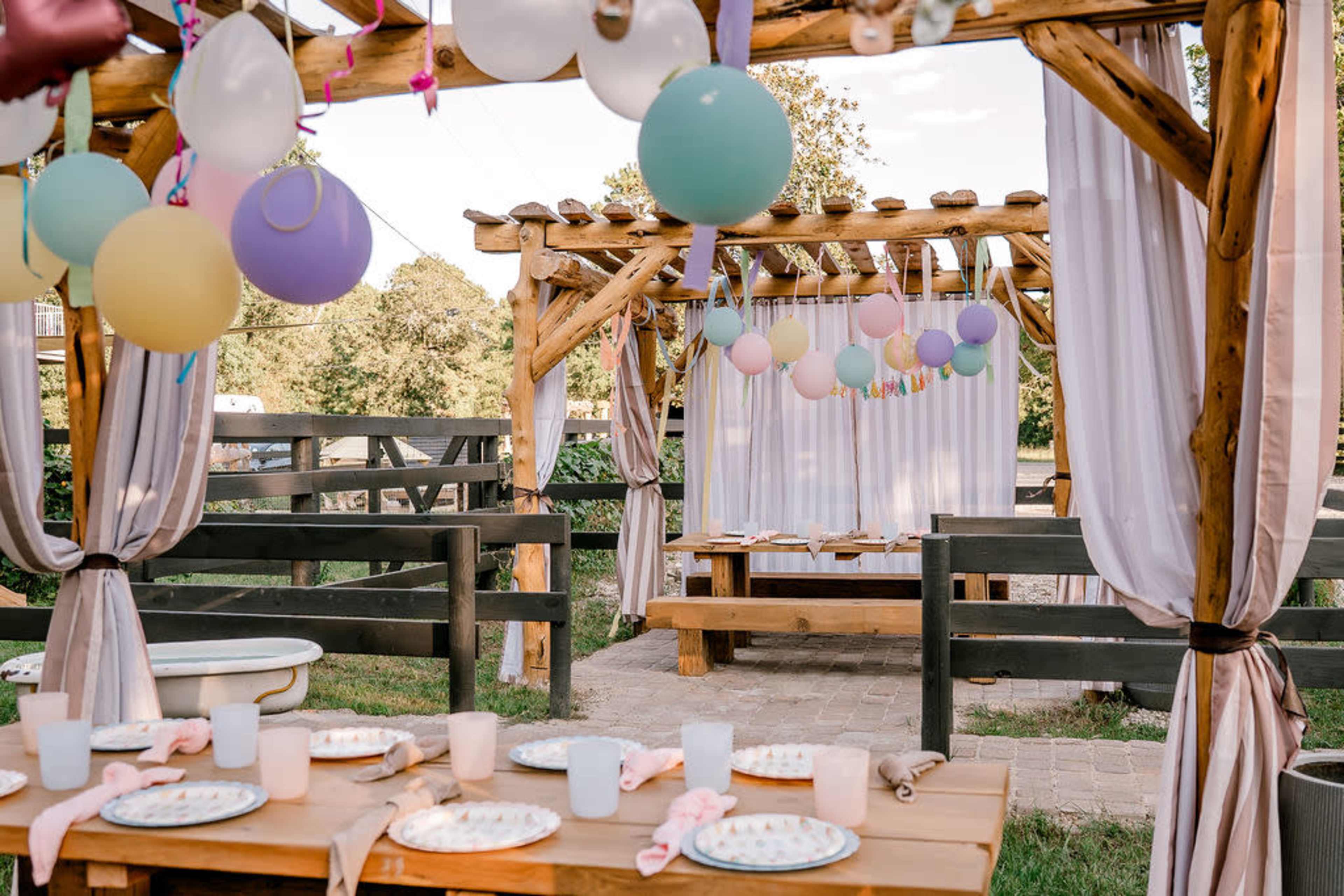 The image depicts a decorated outdoor party space with wooden structures, tables set with plates, and colorful balloons hanging overhead.