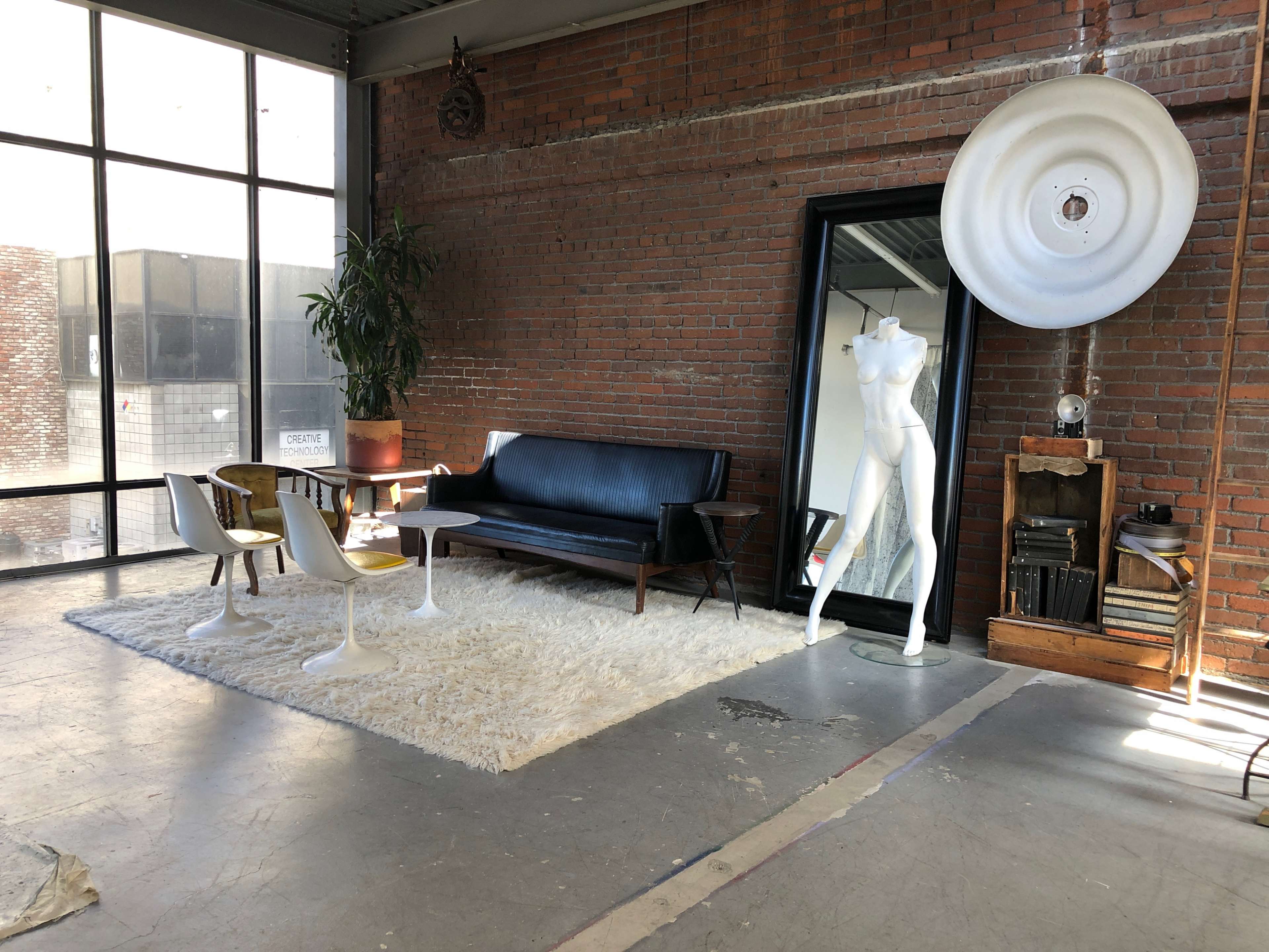 A modern, industrial-style interior featuring a black sofa, a white shag rug, a large mirror, and a mannequin, all against a backdrop of exposed brick walls and large windows.