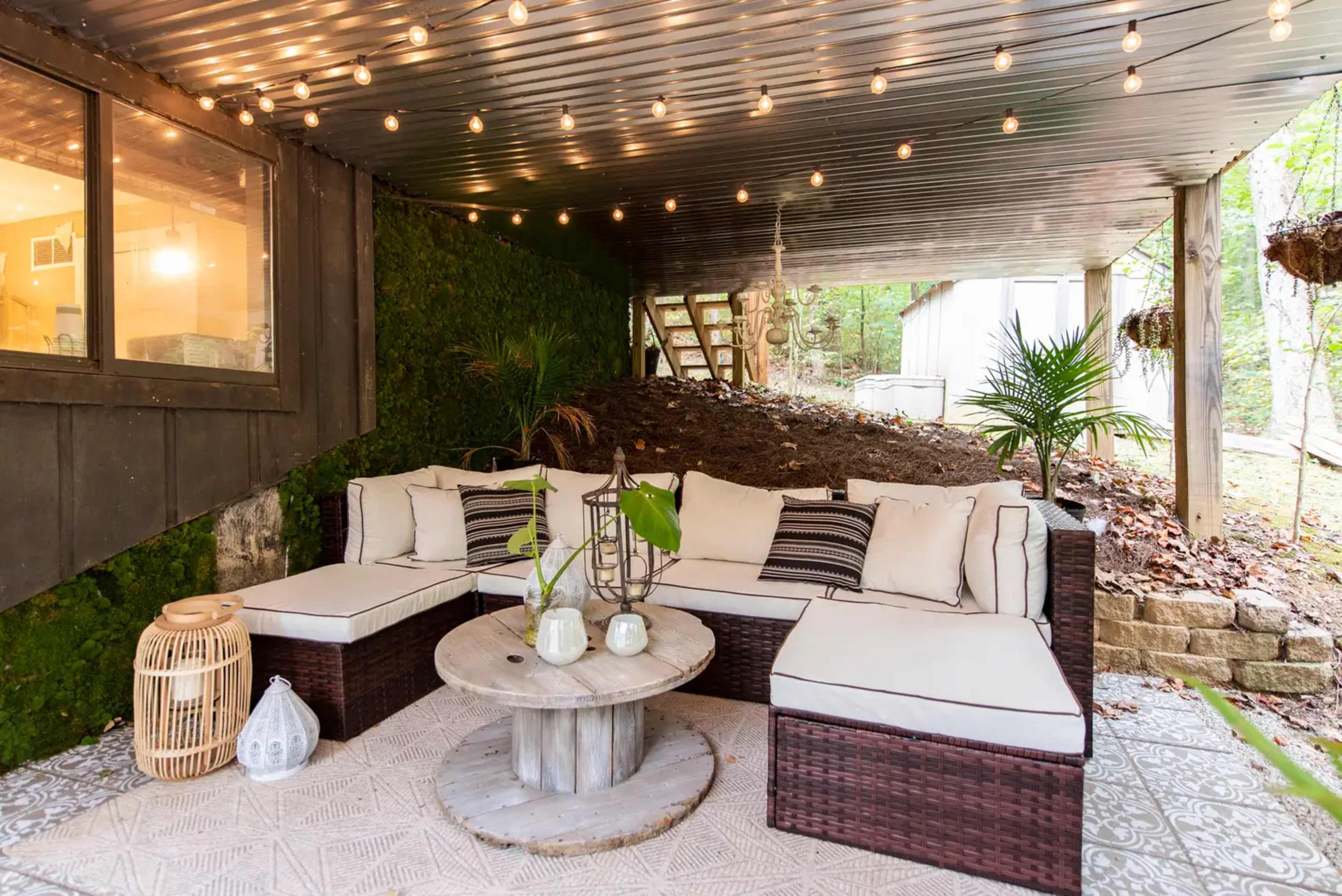 A covered outdoor seating area features a sectional sofa with beige cushions, a round wooden coffee table, and decorative string lights overhead.