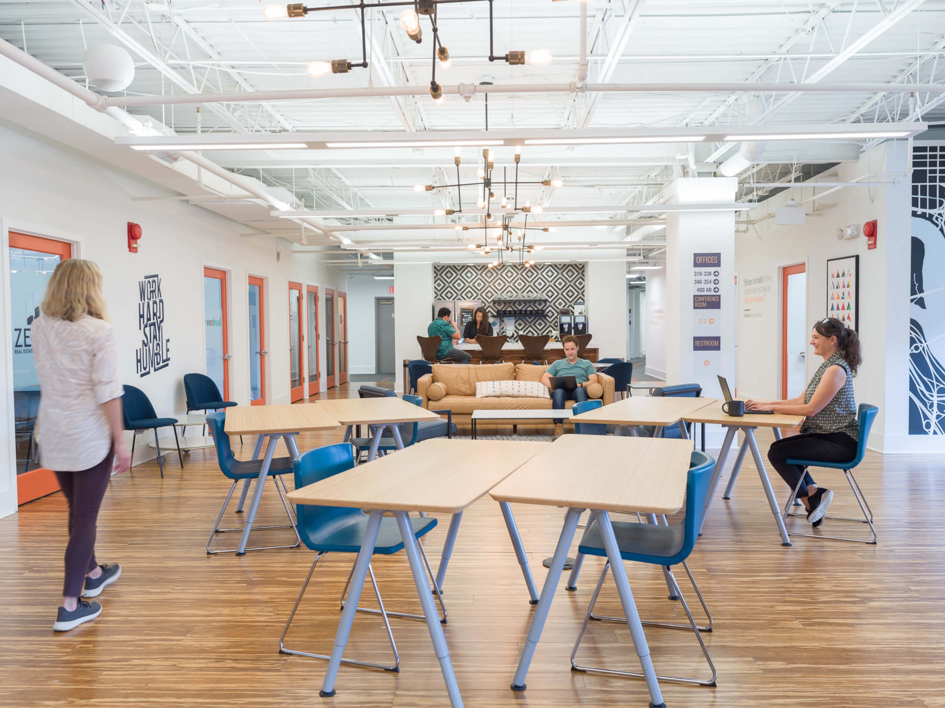 The image shows a modern co-working space with several tables, chairs, and a seating area, inhabited by people engaged in work and conversation.