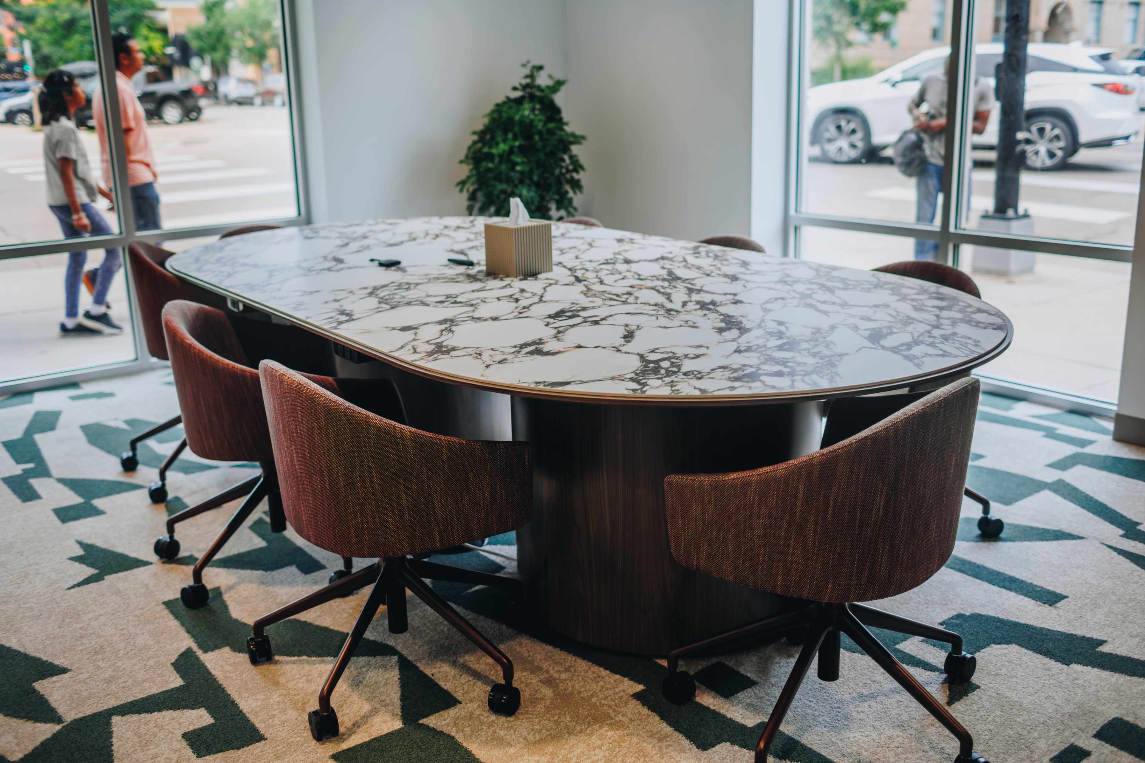 A large, oval conference table with a marble top is surrounded by several rolling chairs in a well-lit room with large windows.