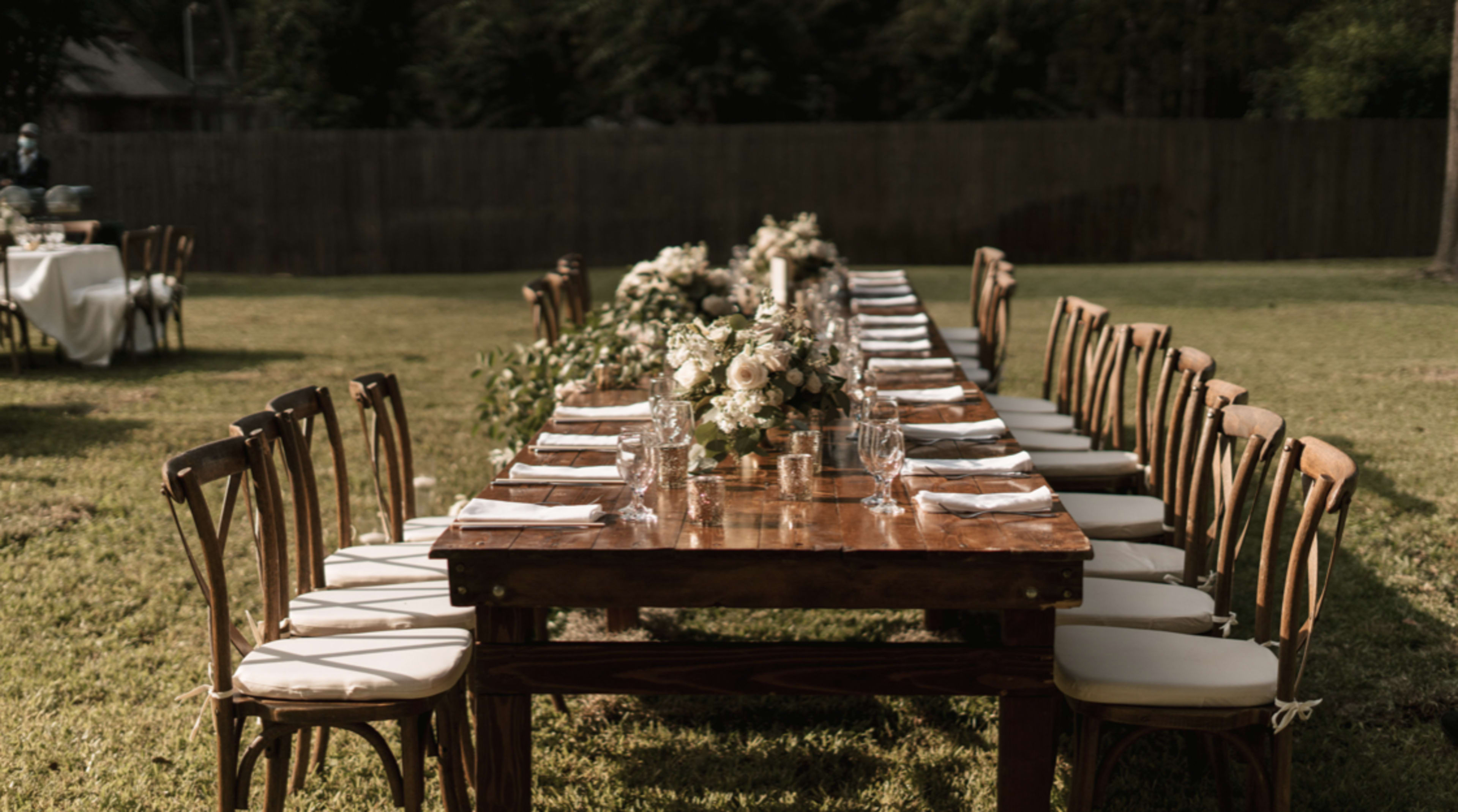 A long wooden dining table is arranged outdoors with neatly placed plates, glasses, and floral centerpieces.