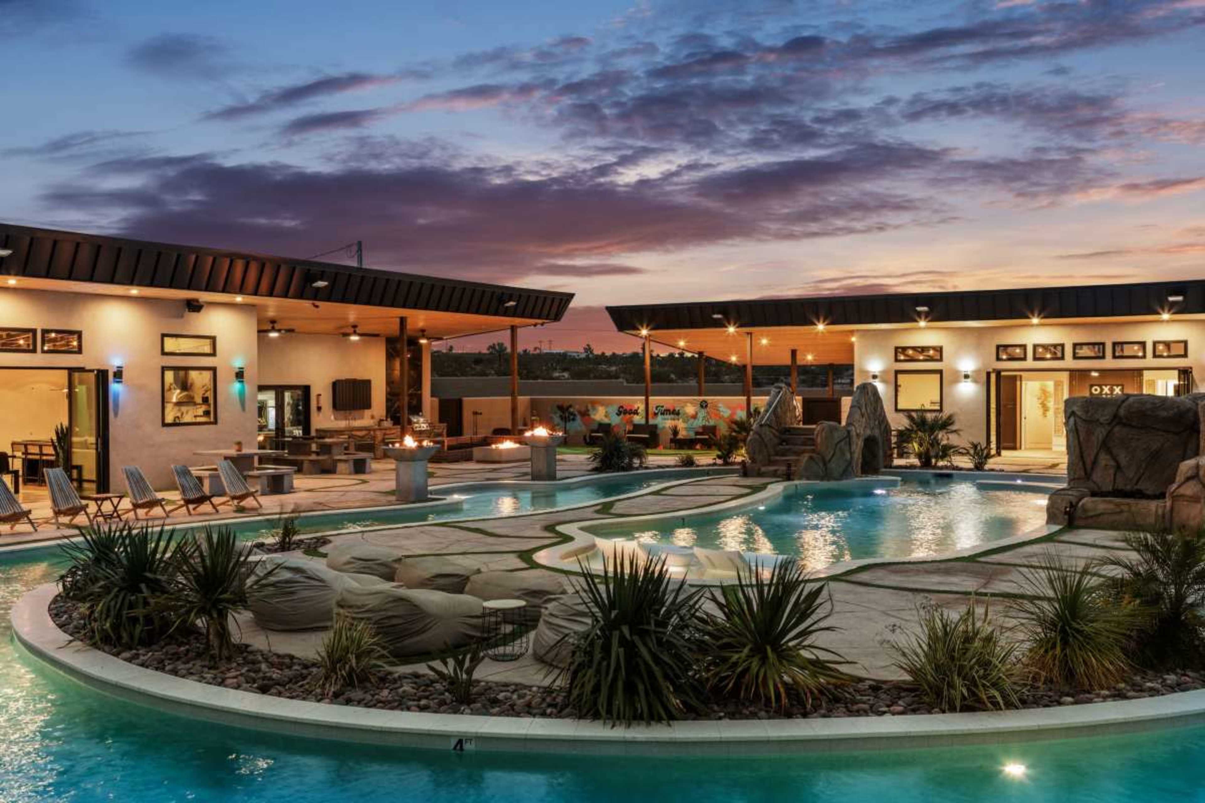 The image shows a spacious outdoor pool area at dusk, featuring a curved pool, rock formations, lounge chairs, and a nearby building with multiple outdoor amenities.