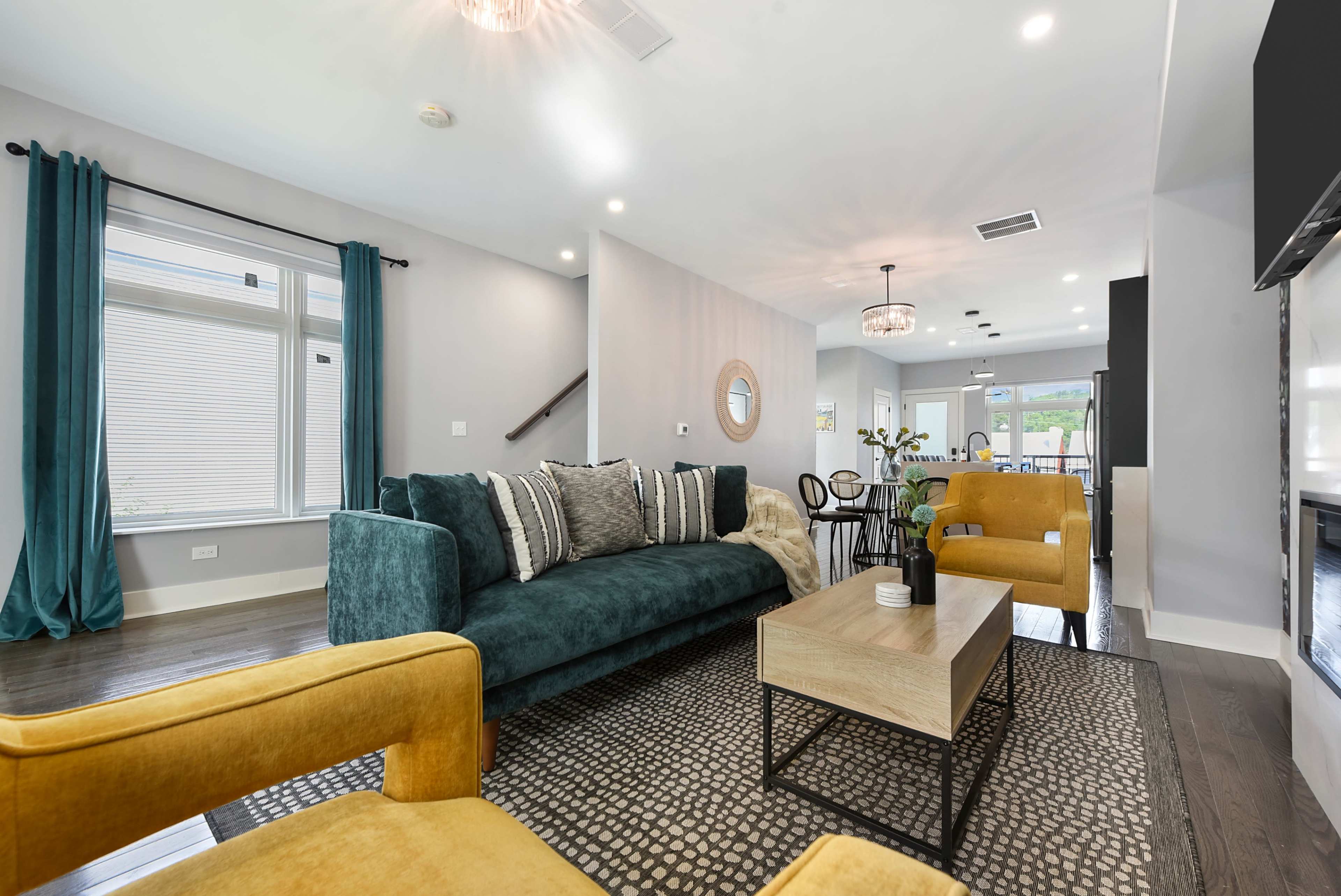 The living room features a green sofa, yellow chairs, a wooden coffee table, and large windows with teal curtains.
