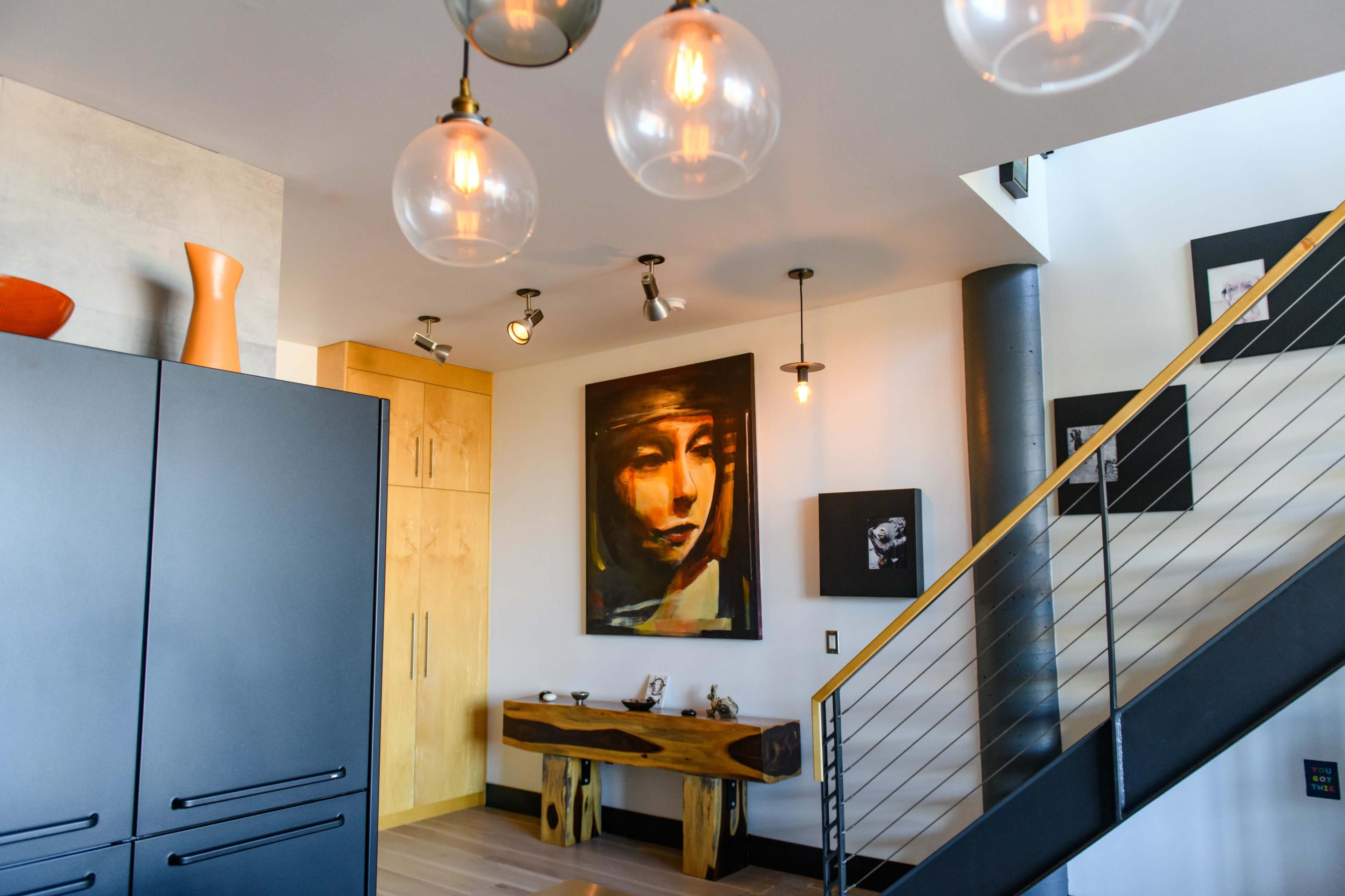 The image features a modern interior space with a staircase, a large painting of a woman on the wall, wooden furniture, and various light fixtures hanging from the ceiling.
