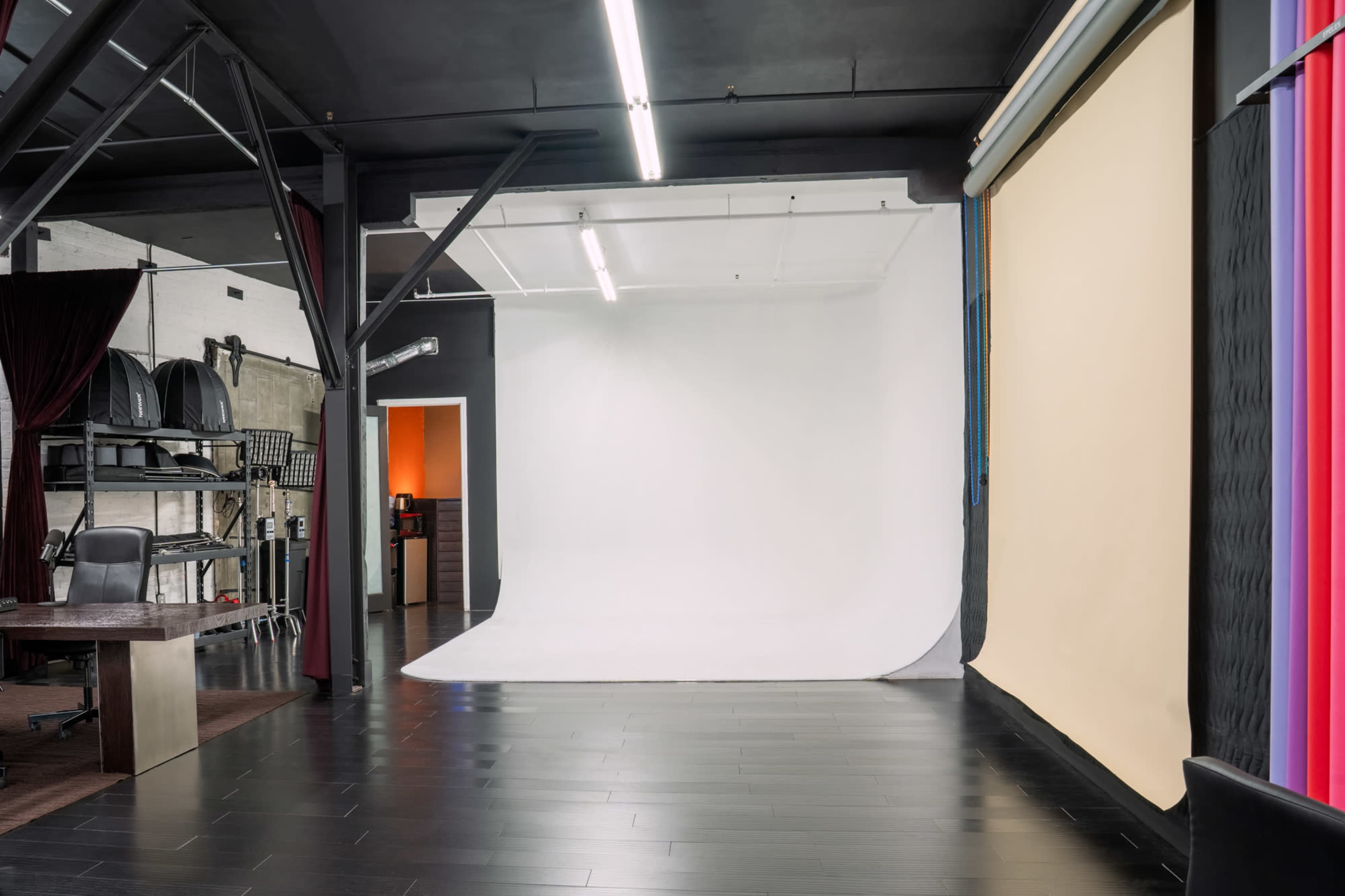 The image shows a photography studio with a large white backdrop and a beige backdrop, surrounded by dark wooden flooring and studio equipment.