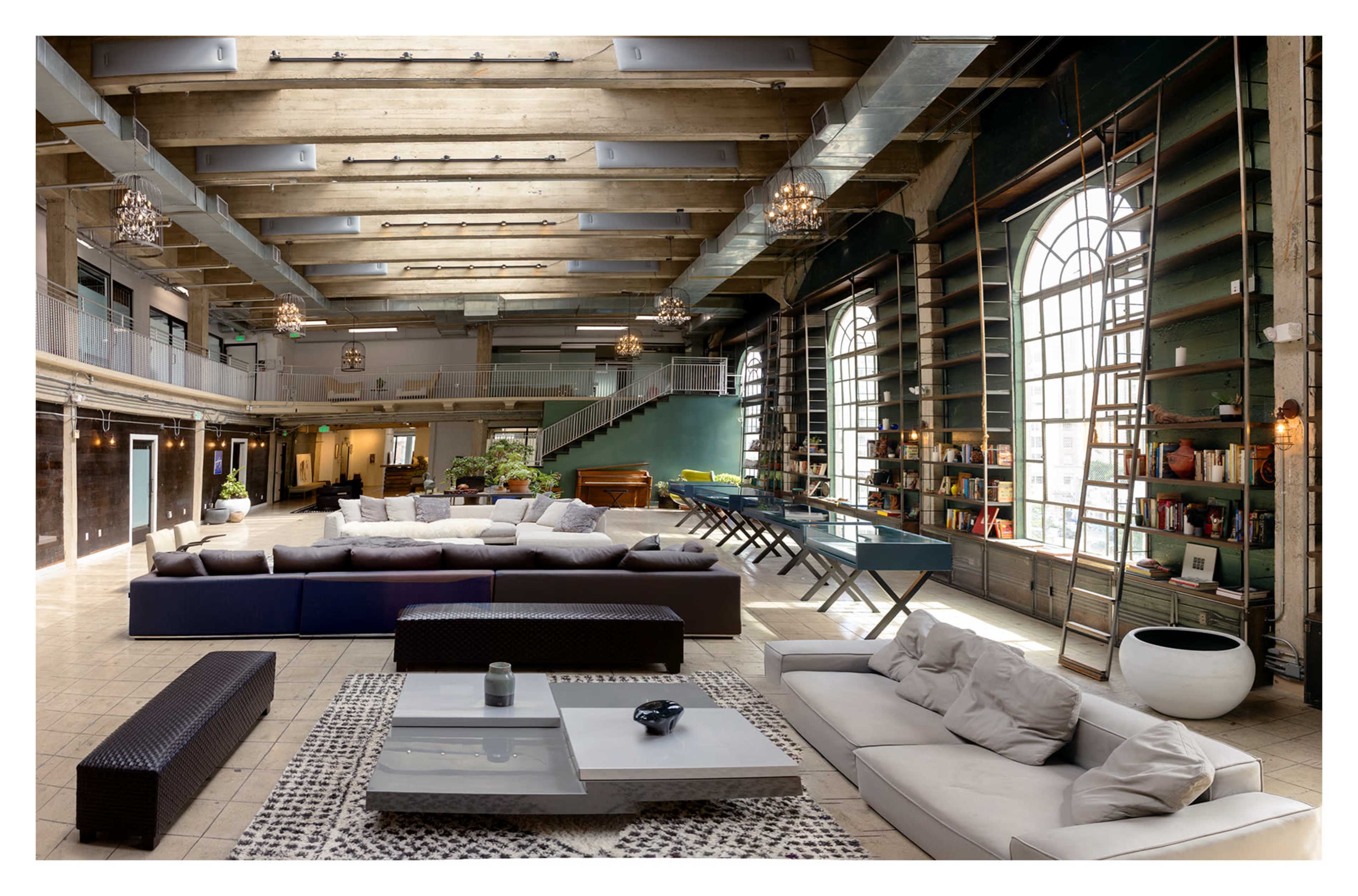 The image shows a spacious modern living area with high ceilings, large windows, and various seating arrangements, including a sectional sofa, side tables, and a dining area in the background.