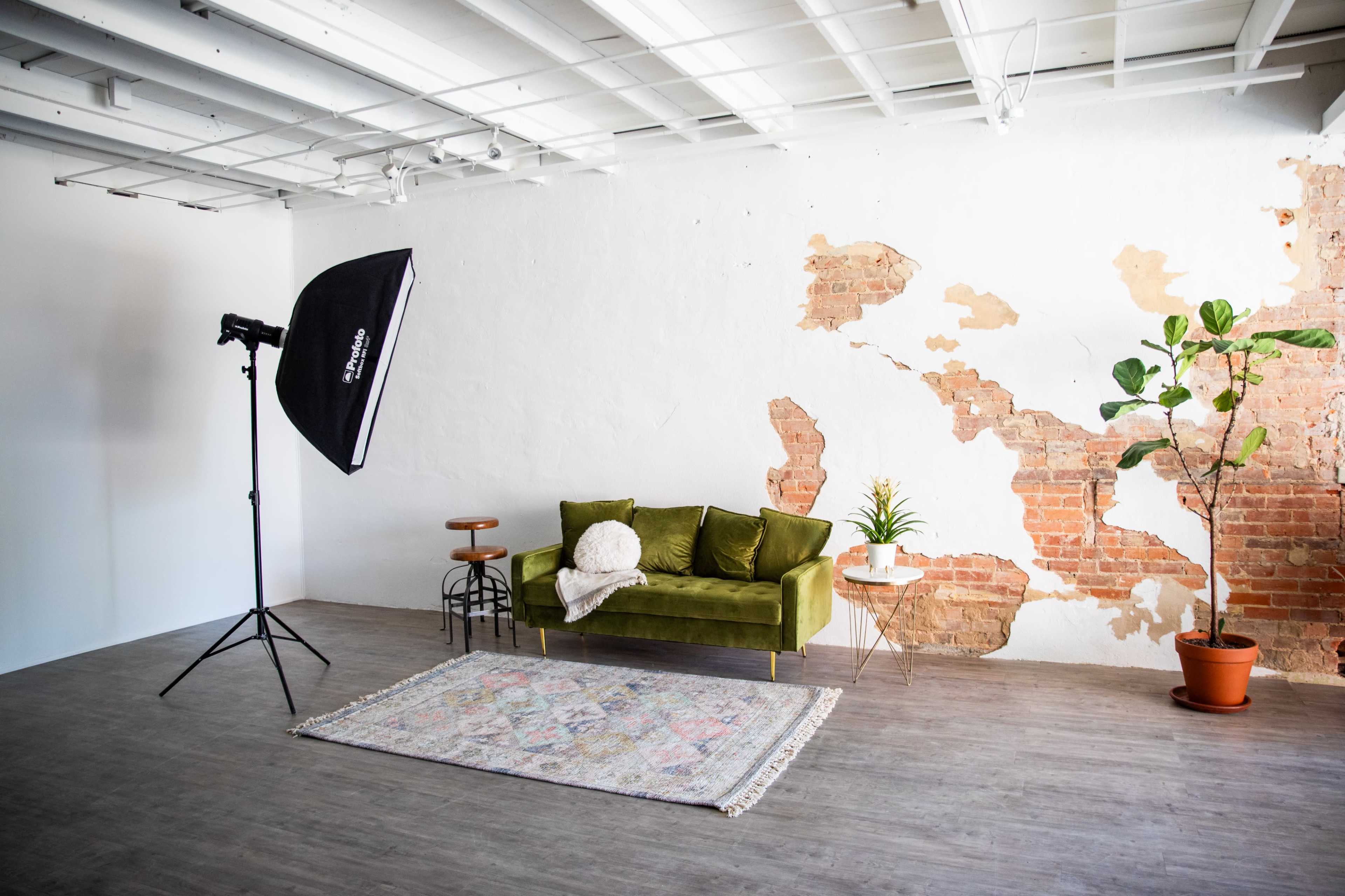 The scene features a green velvet sofa placed against a wall with exposed brick, accompanied by a photography light, a small side table with a plant, and a patterned rug on the wooden floor.