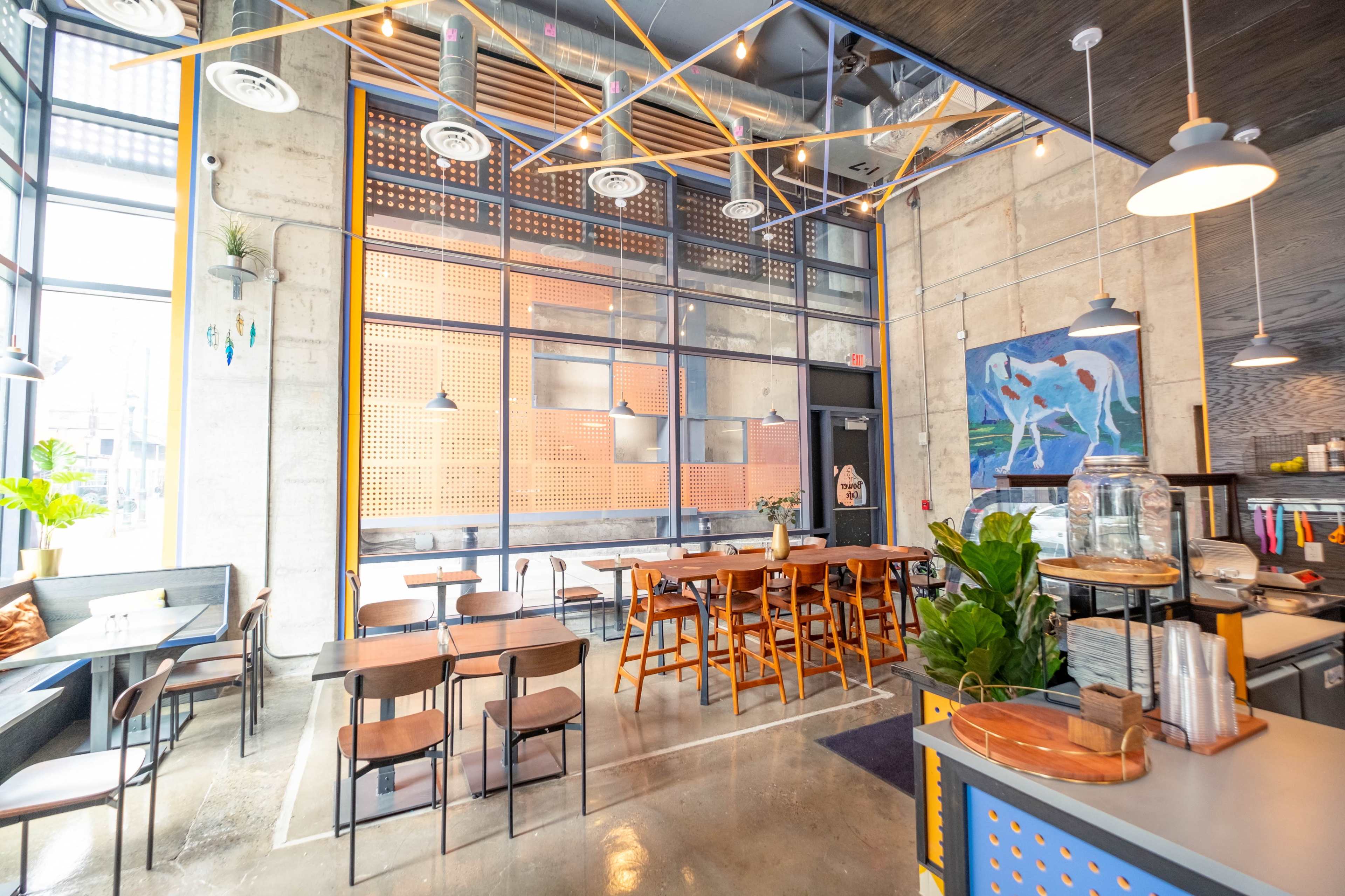 A modern cafe interior with metal and wooden furniture, large windows, and a colorful wall art piece.