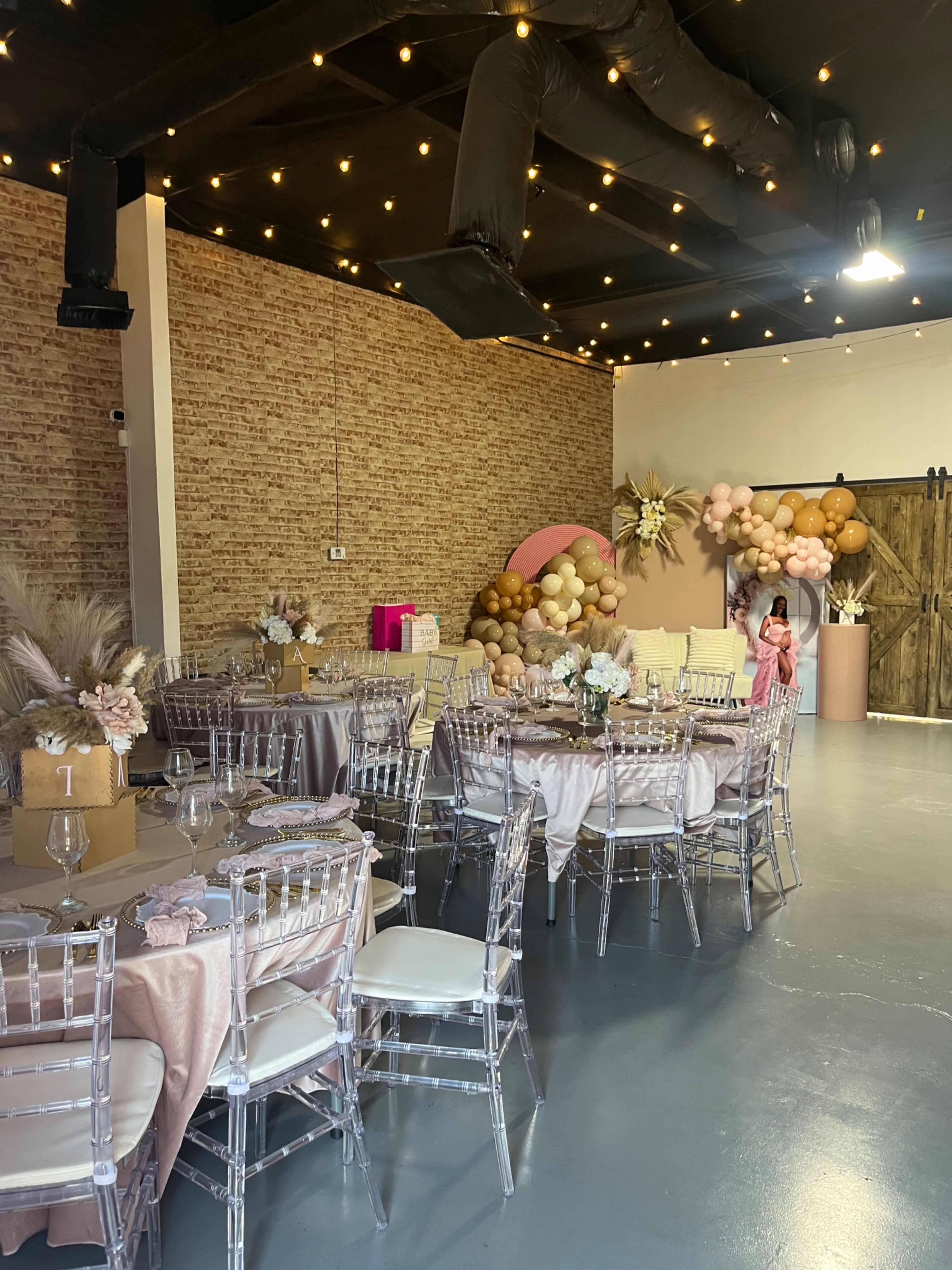 A decorated event space features several round tables set with floral centerpieces, draped in elegant tablecloths, alongside a backdrop of balloons and decor.