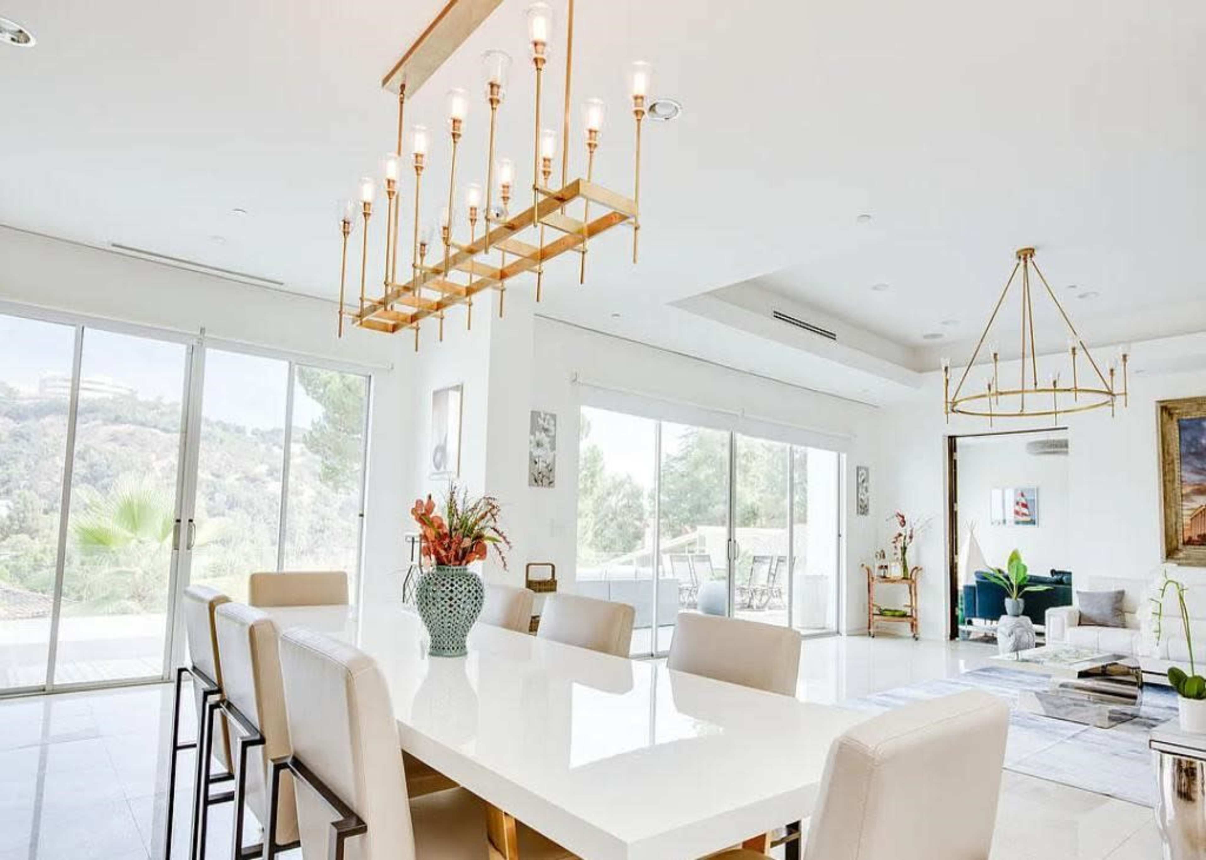The image shows a modern dining area with a long white table surrounded by beige chairs, highlighted by elegant light fixtures overhead and large windows allowing natural light to enter.