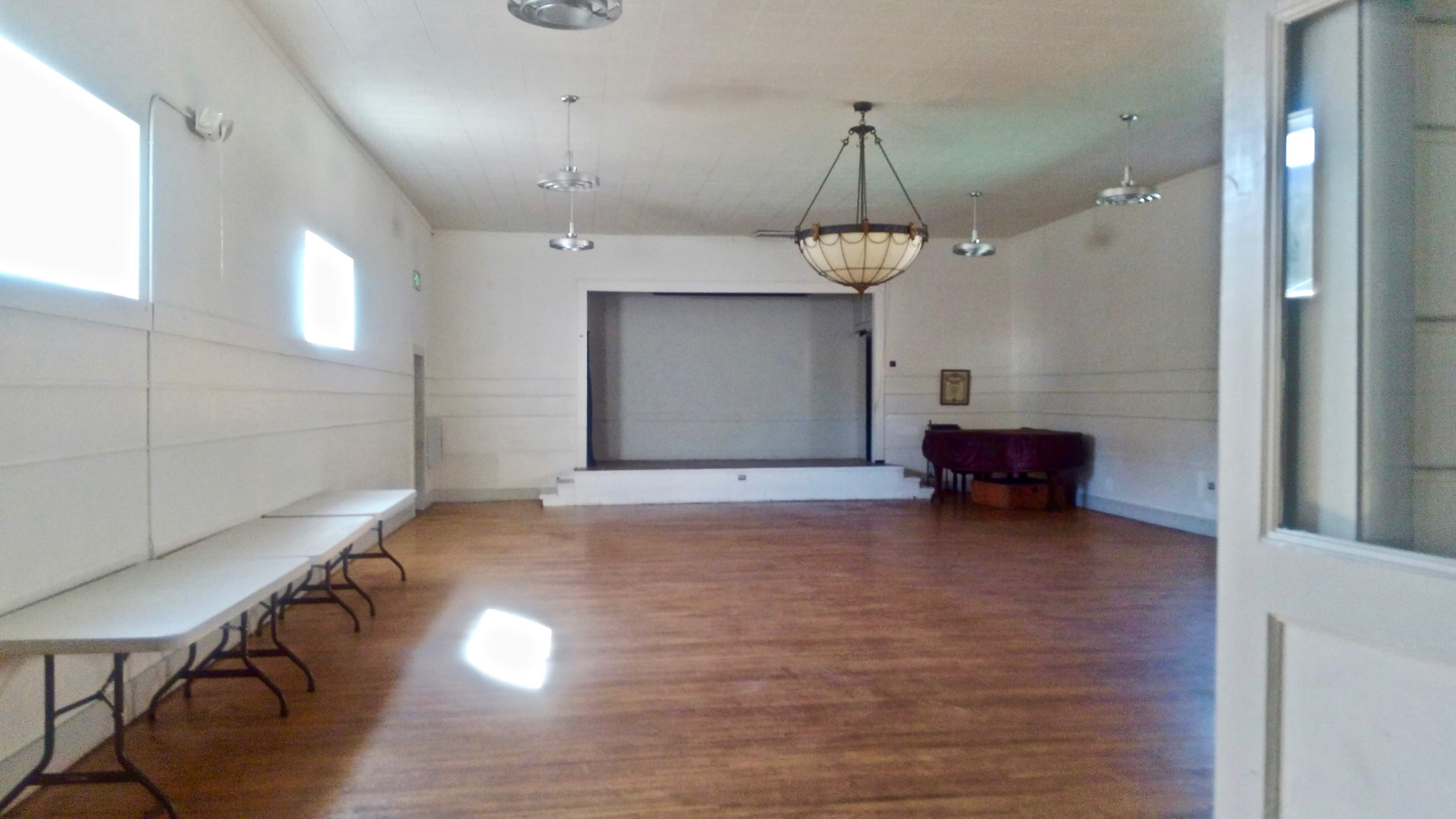 A spacious, empty room with a wooden floor, featuring a stage at the far end and several folding tables along one wall.
