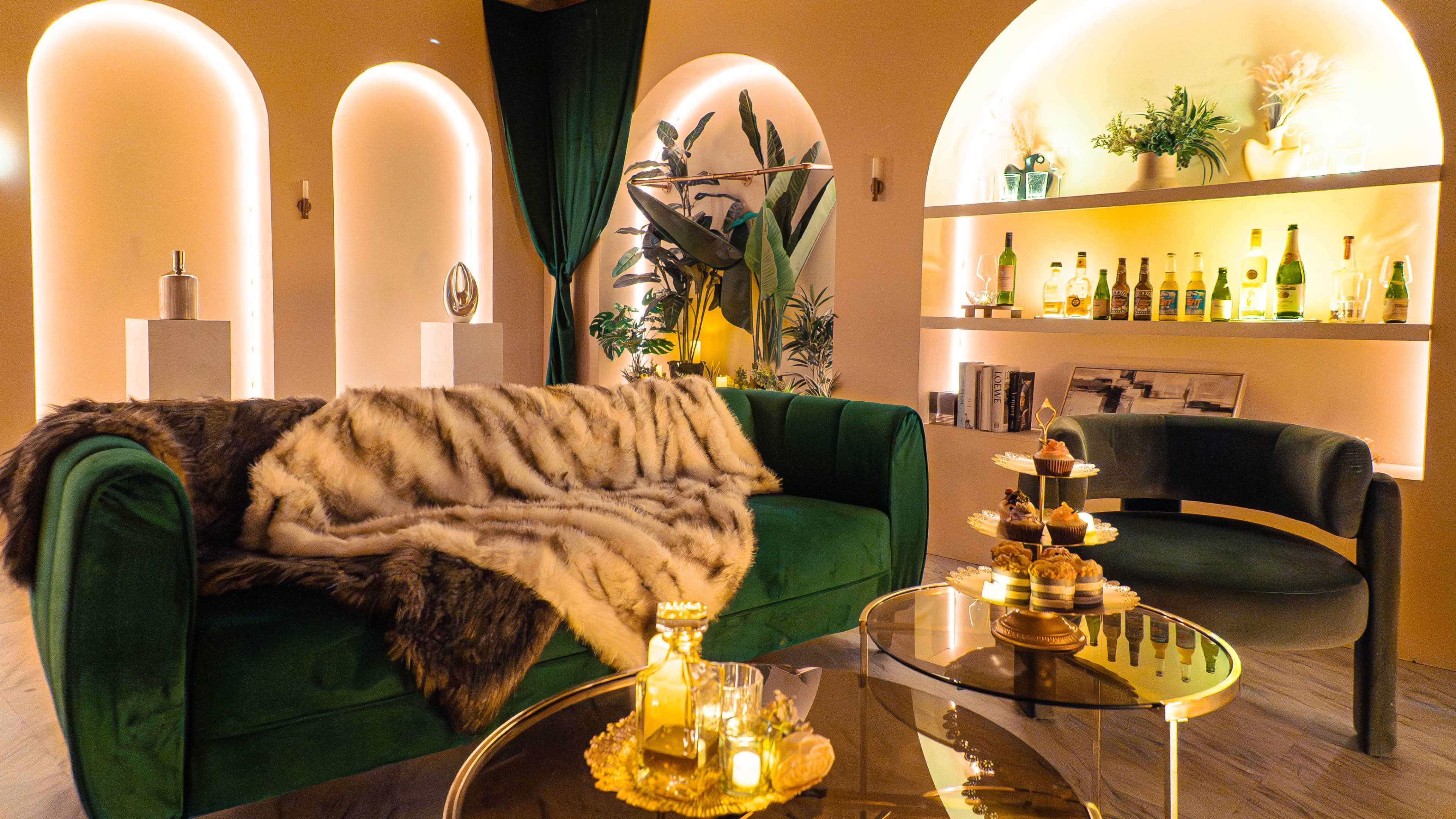 The image depicts a stylish interior featuring a green velvet sofa draped with a fur throw, a glass coffee table, and a shelf showcasing various bottles and decorative items, all illuminated by warm lighting.