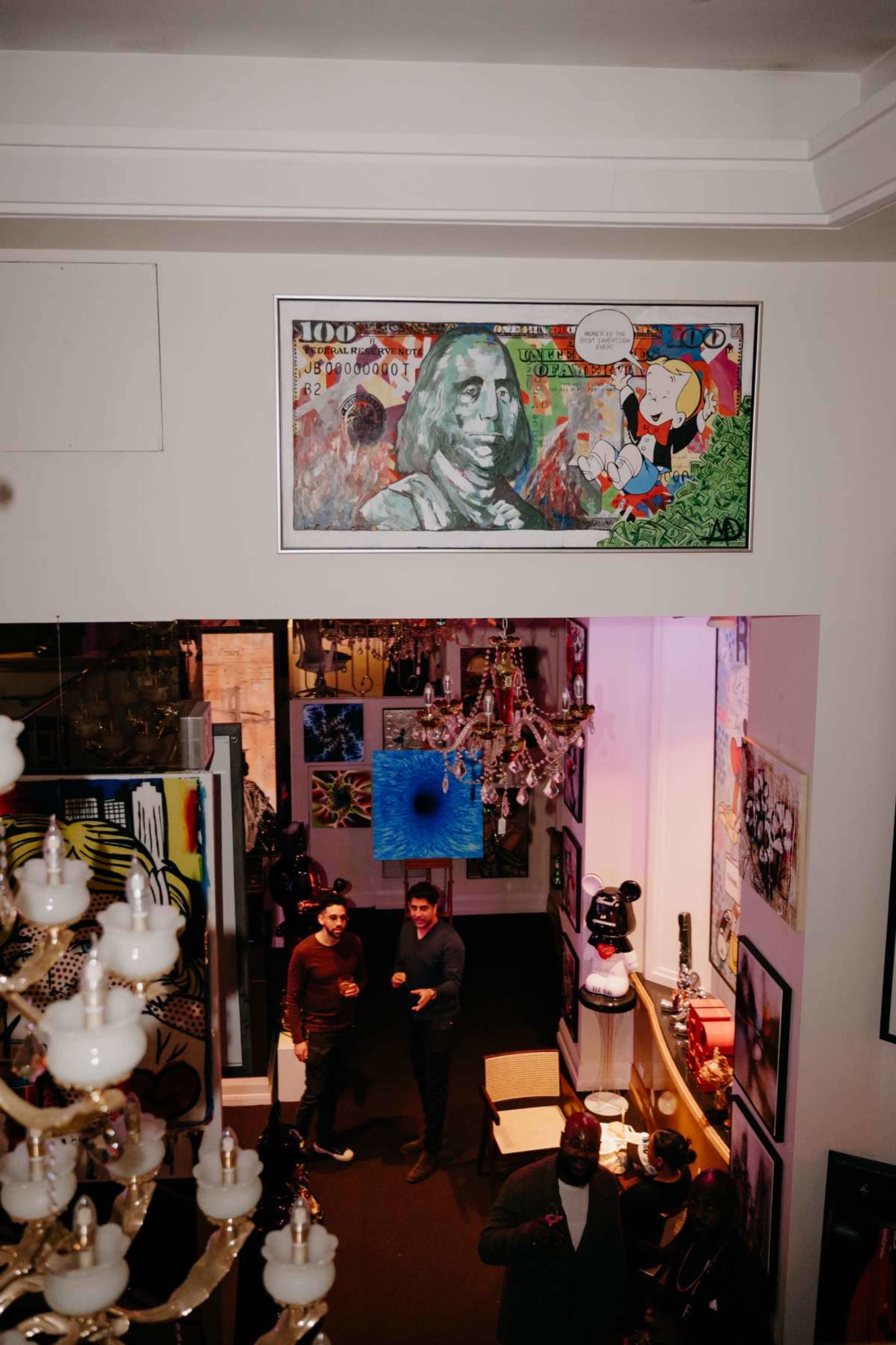 The image shows a colorful art gallery with various artworks displayed on the walls, including a prominent piece featuring a large bill with a portrait, while two people are standing and talking in the center.