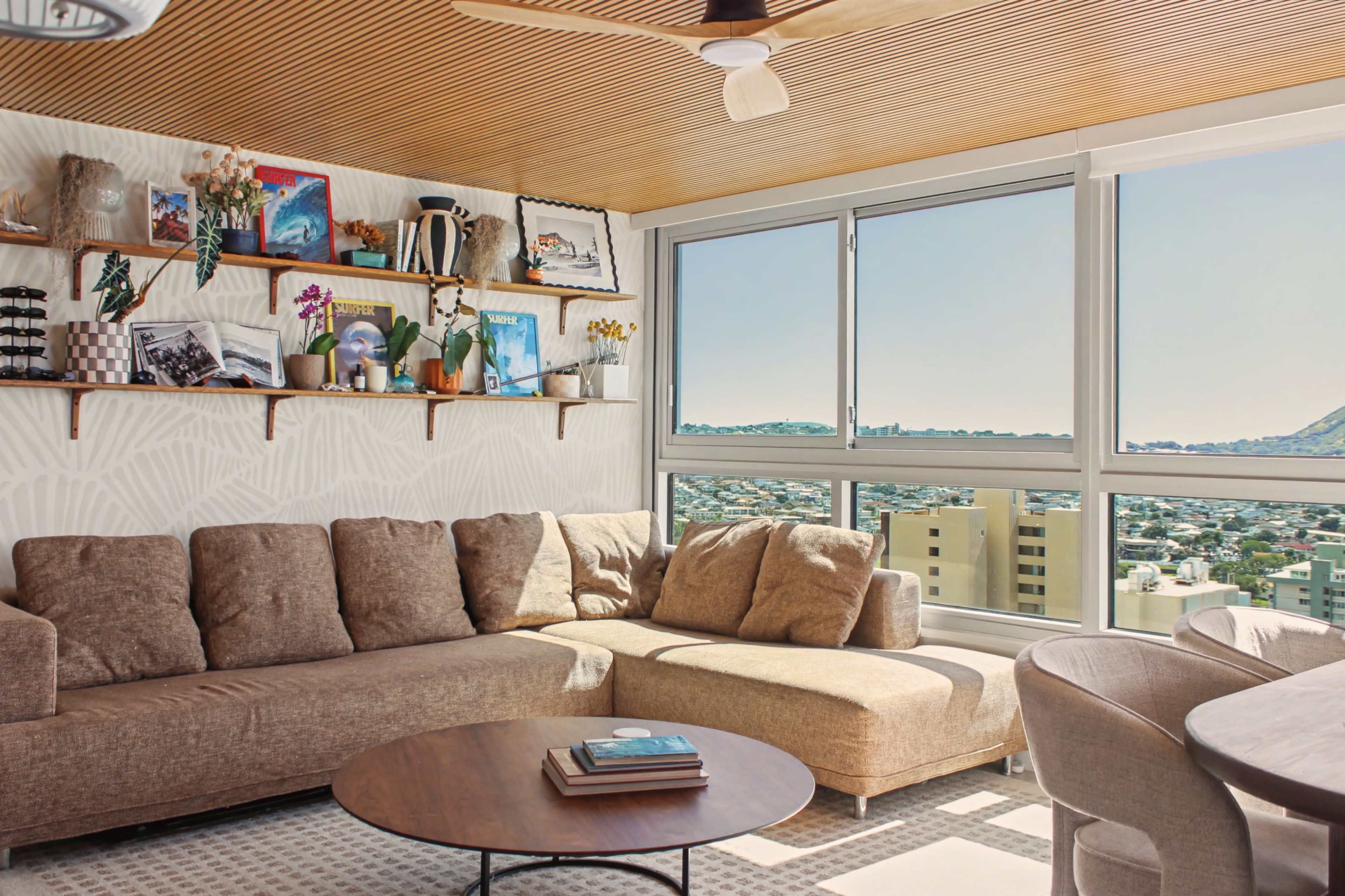 The image shows a cozy living room with a large brown sectional sofa, a round coffee table, and a wall shelf filled with art and plants, all framed by a large window showcasing a city view.