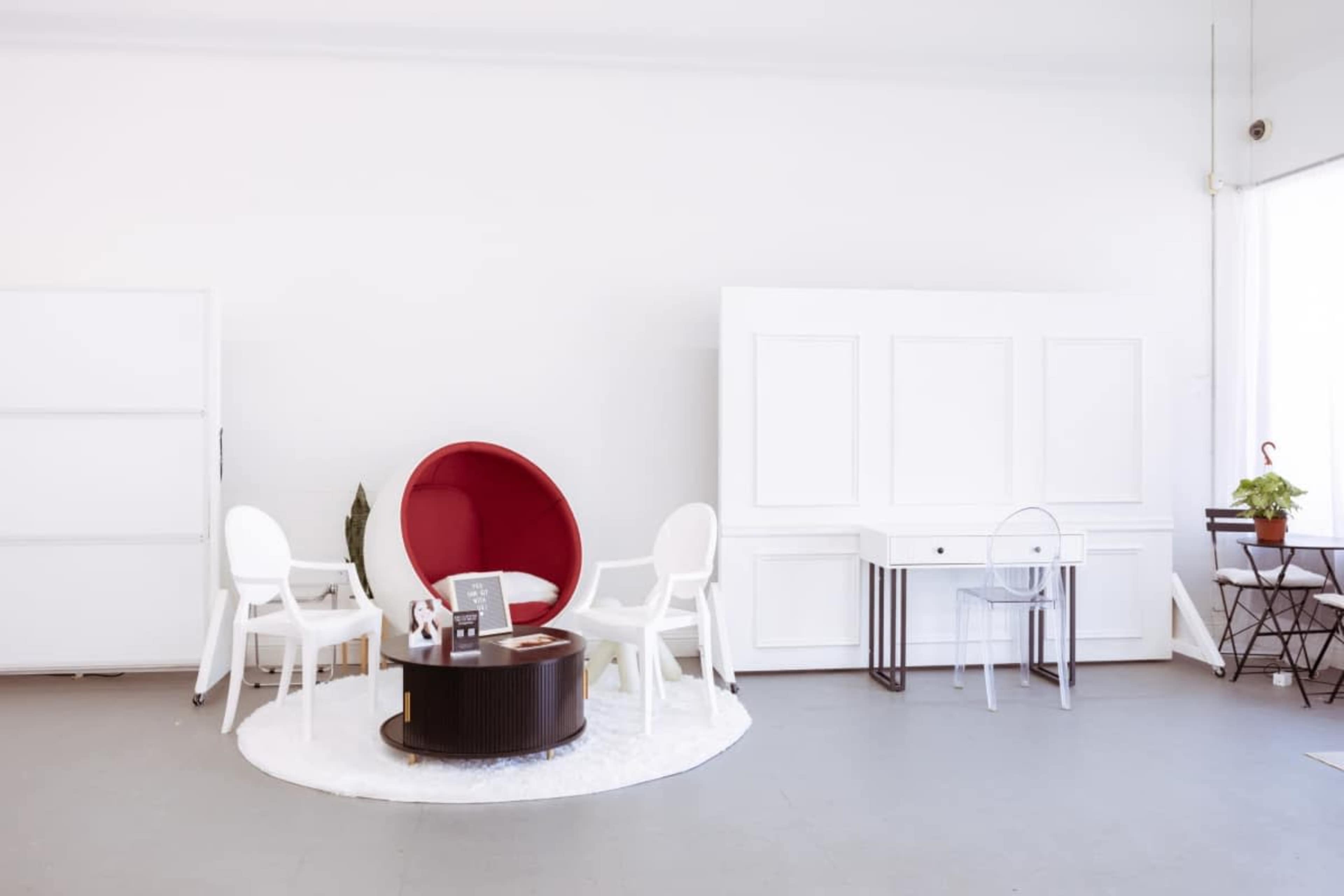 The image shows a minimalist interior with a red modern chair, white accent chairs, a round coffee table, and a small desk, all set against a light-colored background.