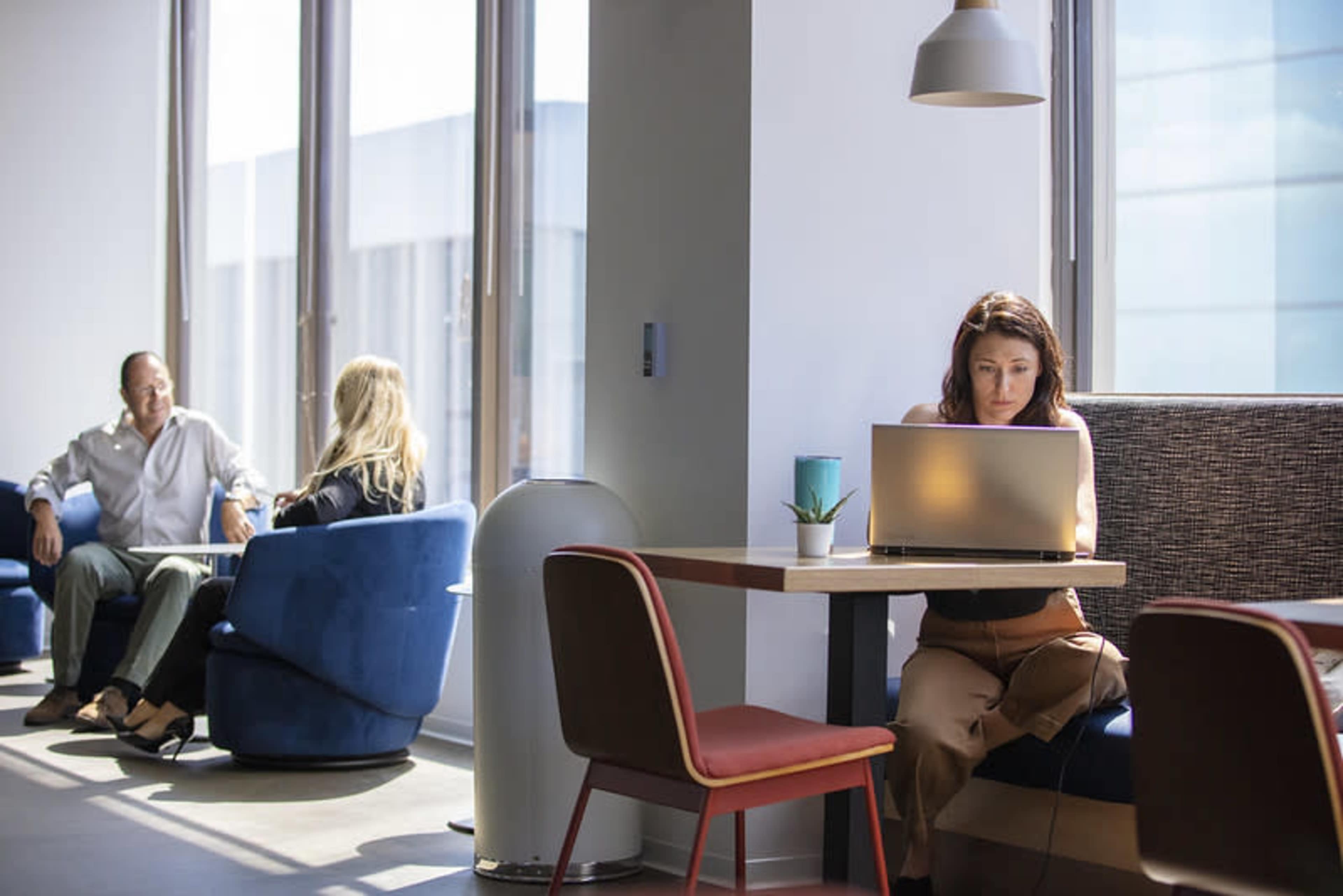 A woman works on a laptop at a table while two people converse in upholstered chairs nearby in a modern, well-lit space.