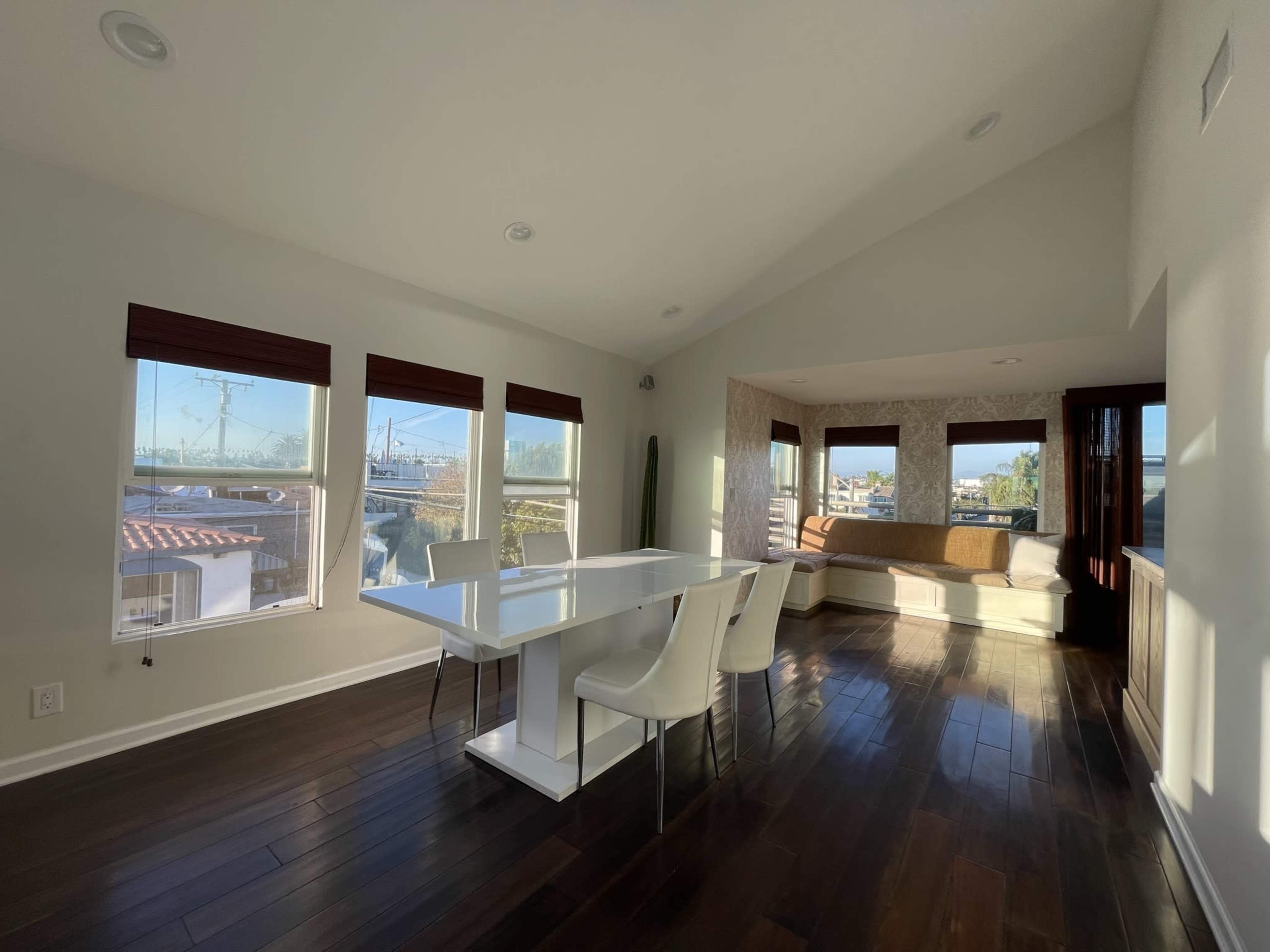 A modern dining area features a white table with four chairs, large windows allowing natural light, and a cozy seating nook in the corner.