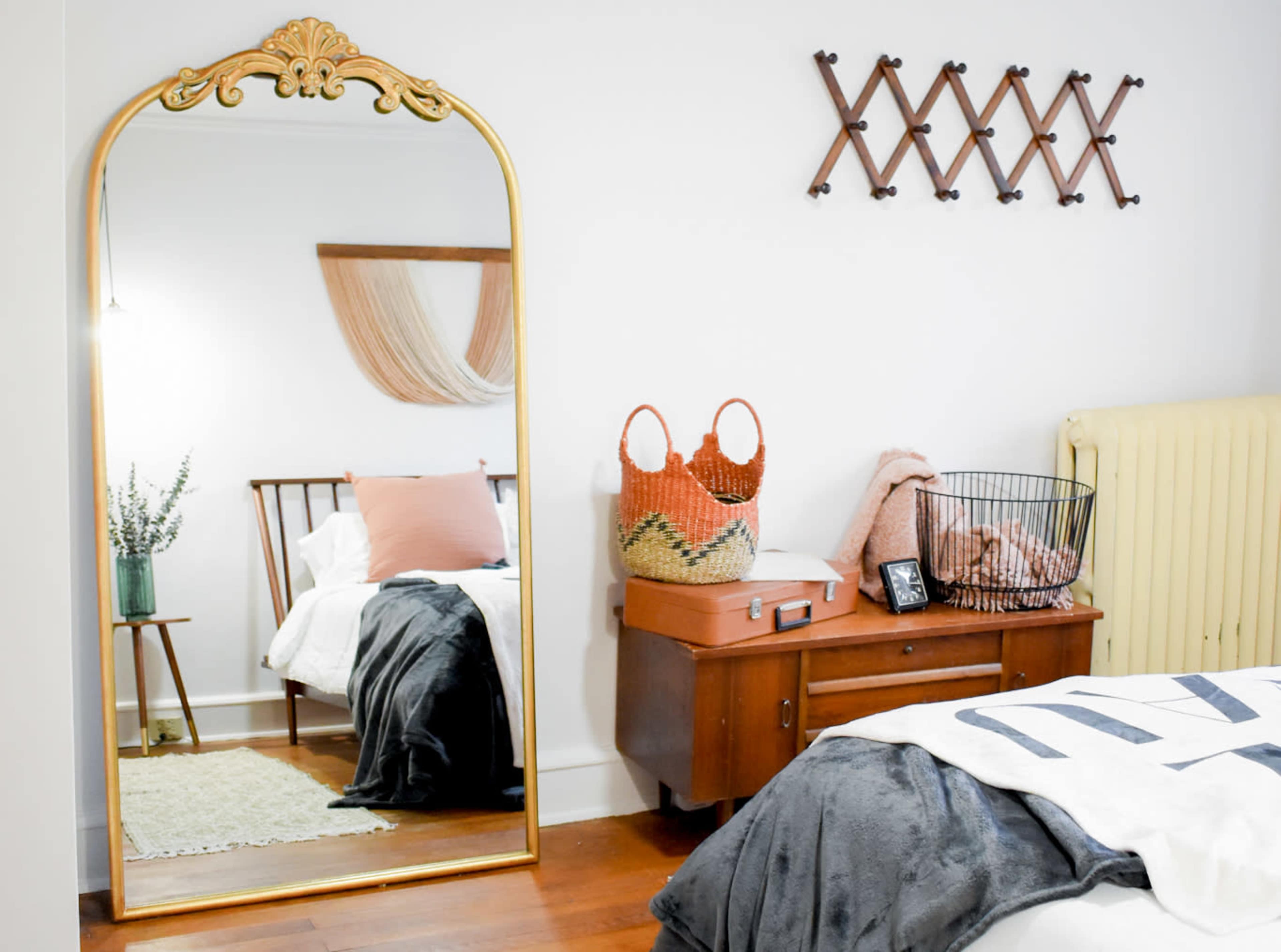 A bedroom featuring a large gold-framed mirror, a wooden bed with decorative pillows, a woven basket, and a vintage-style chest of drawers.