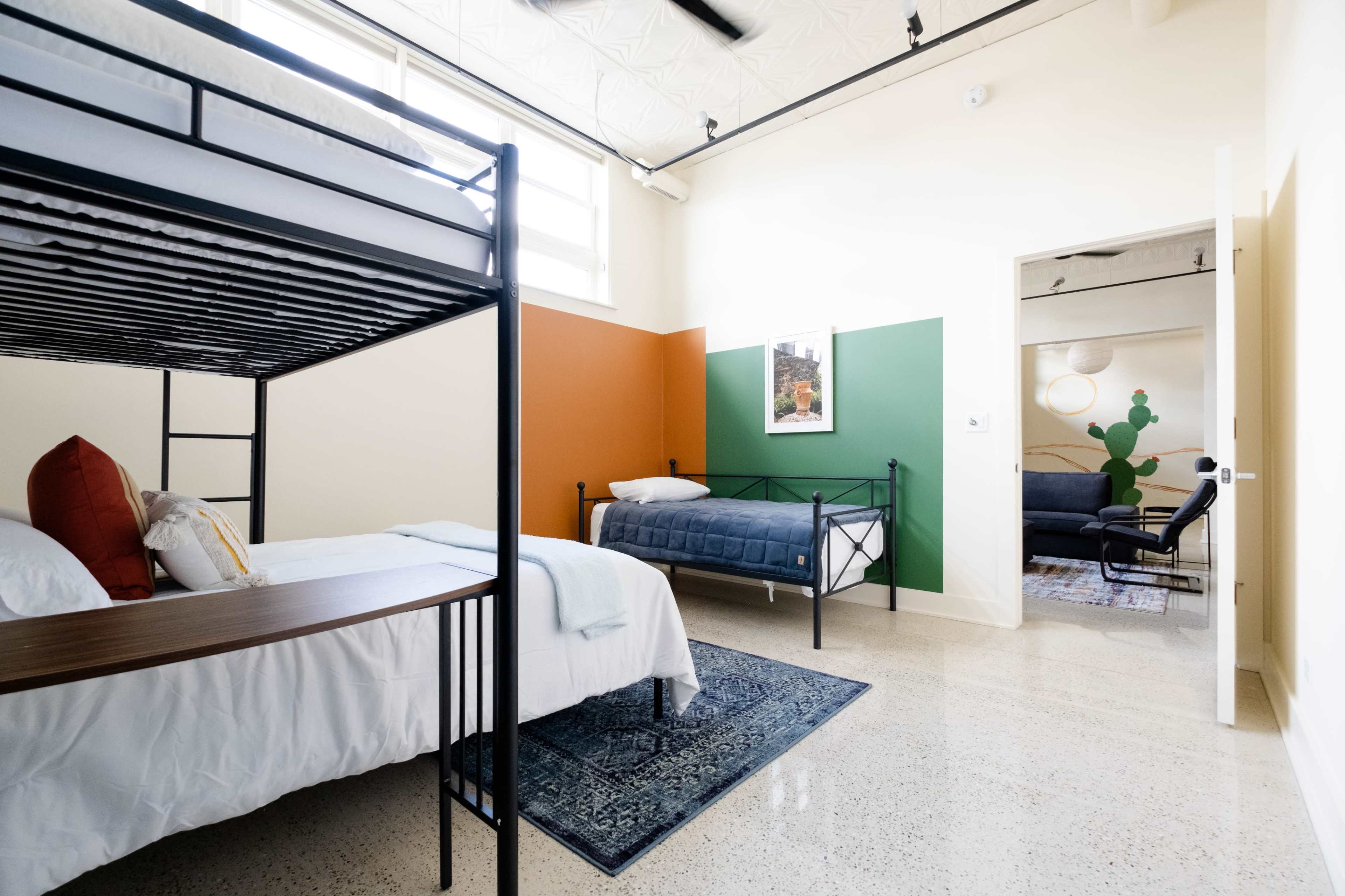 A modern bedroom with a bunk bed and a single bed, featuring a colorful wall with green and orange sections, and a seating area in the background.